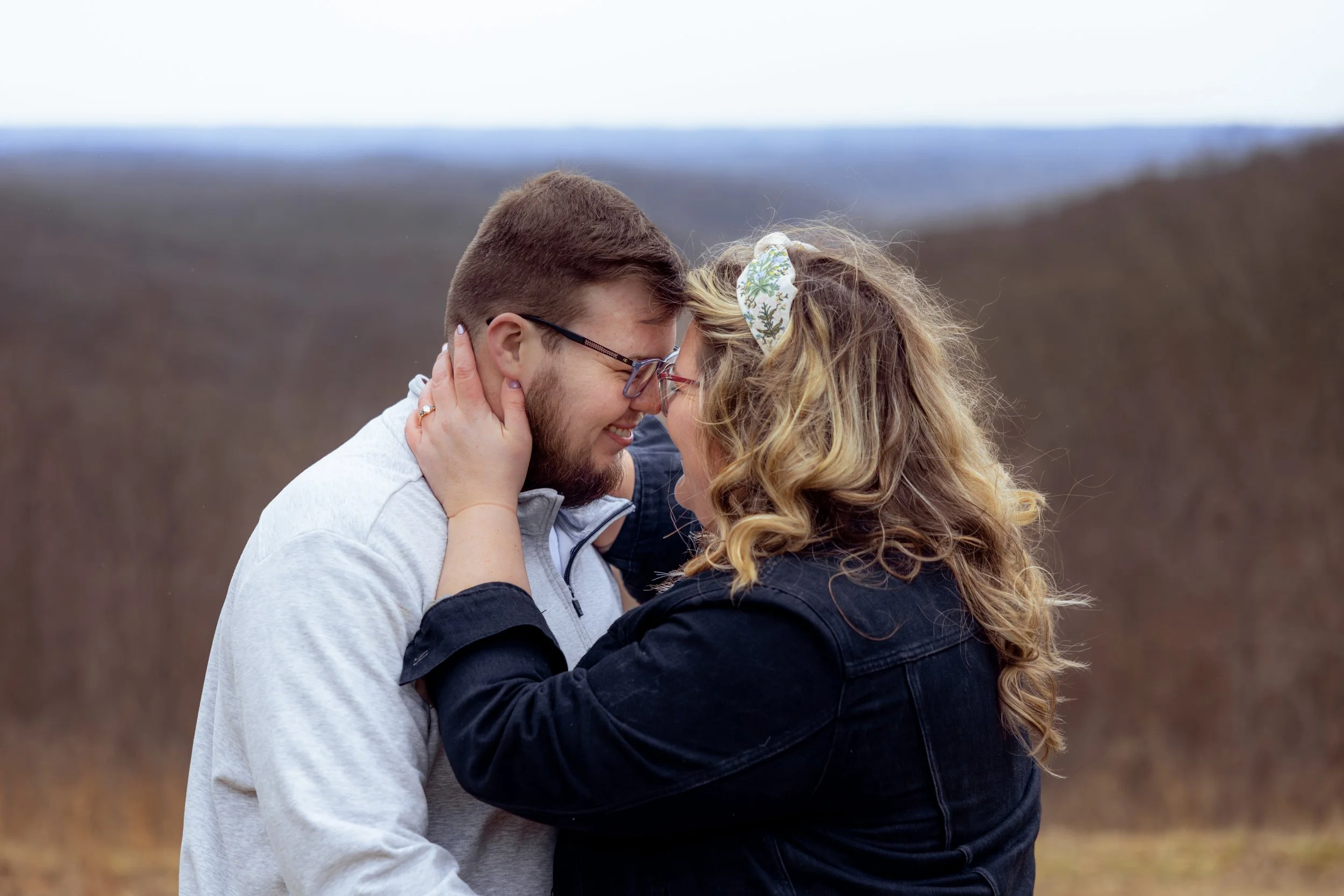 A couple touching foreheads and smiling outdoors during daytime after getting engaged, with the woman wearing her engagement ring.