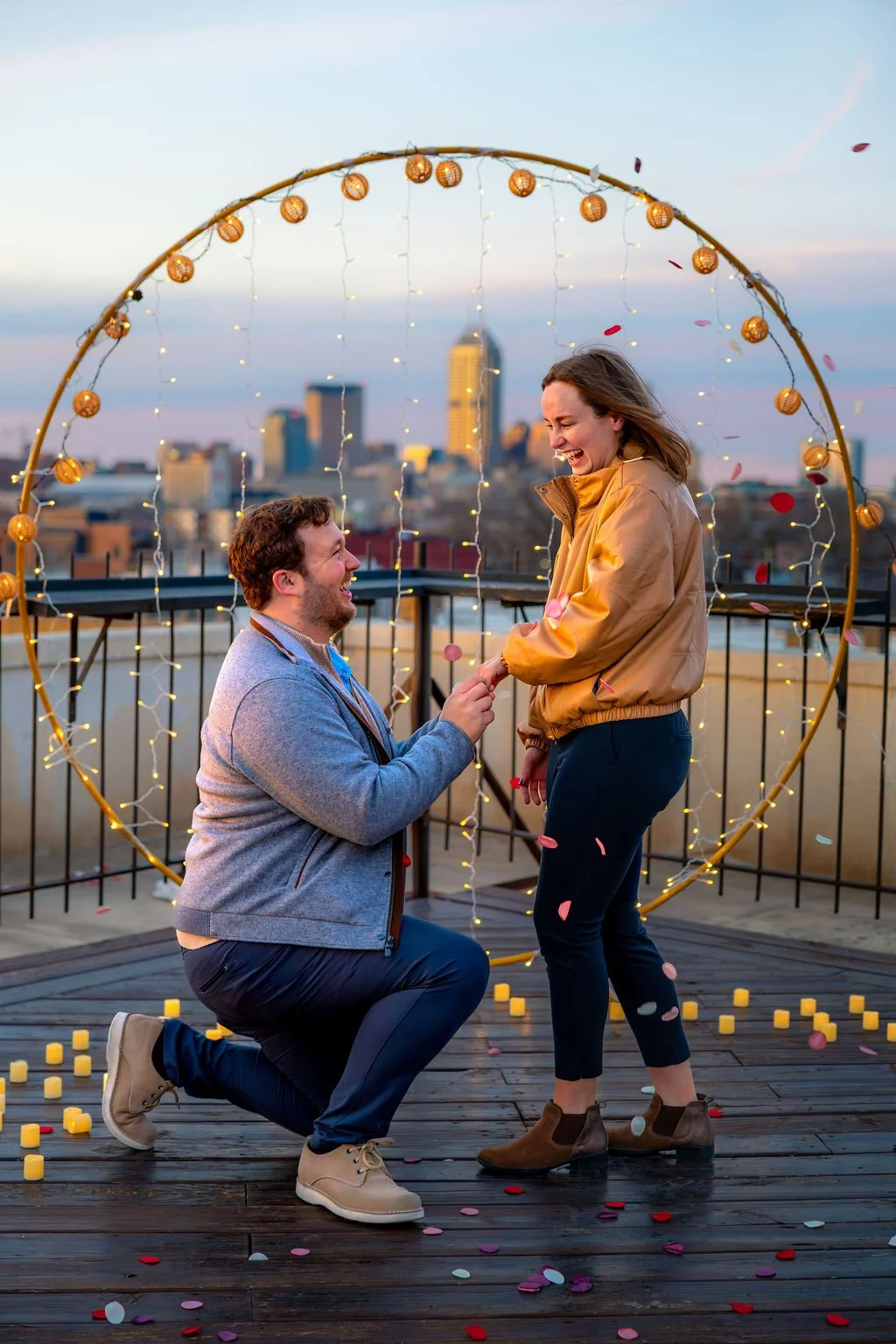 A man proposing marriage to a woman on a rooftop with downtown Indianapolis in the background, surrounded by lights and confetti.