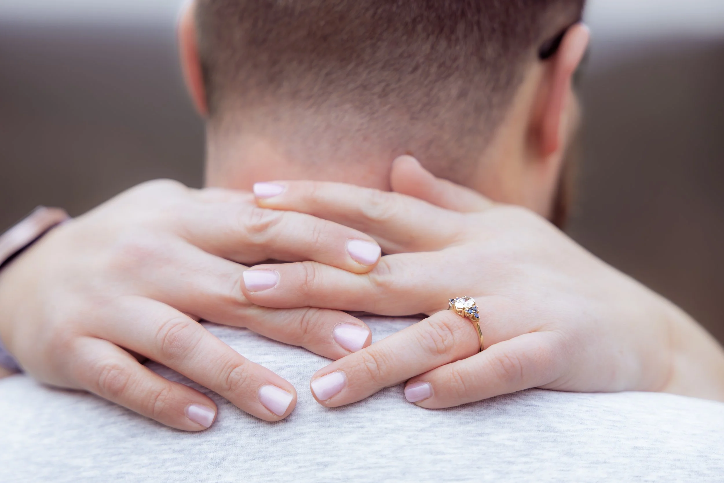 A woman hugging a man from behind, her hands resting on his shoulders. She wears a ring with a large gemstone on her finger.