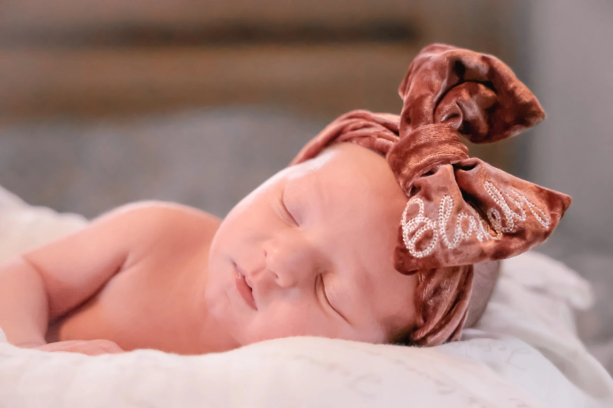 Close-up of a sleeping newborn baby wearing a large, brown satin bow headband with embroidered text, lying on a soft, white blanket.