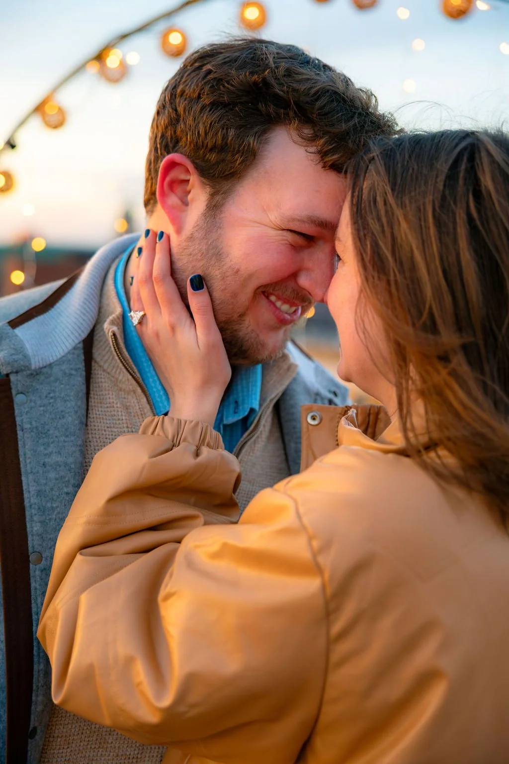 A couple smiling and touching foreheads, with the woman’s hand on the man’s cheek, outdoor setting with lights in the background.