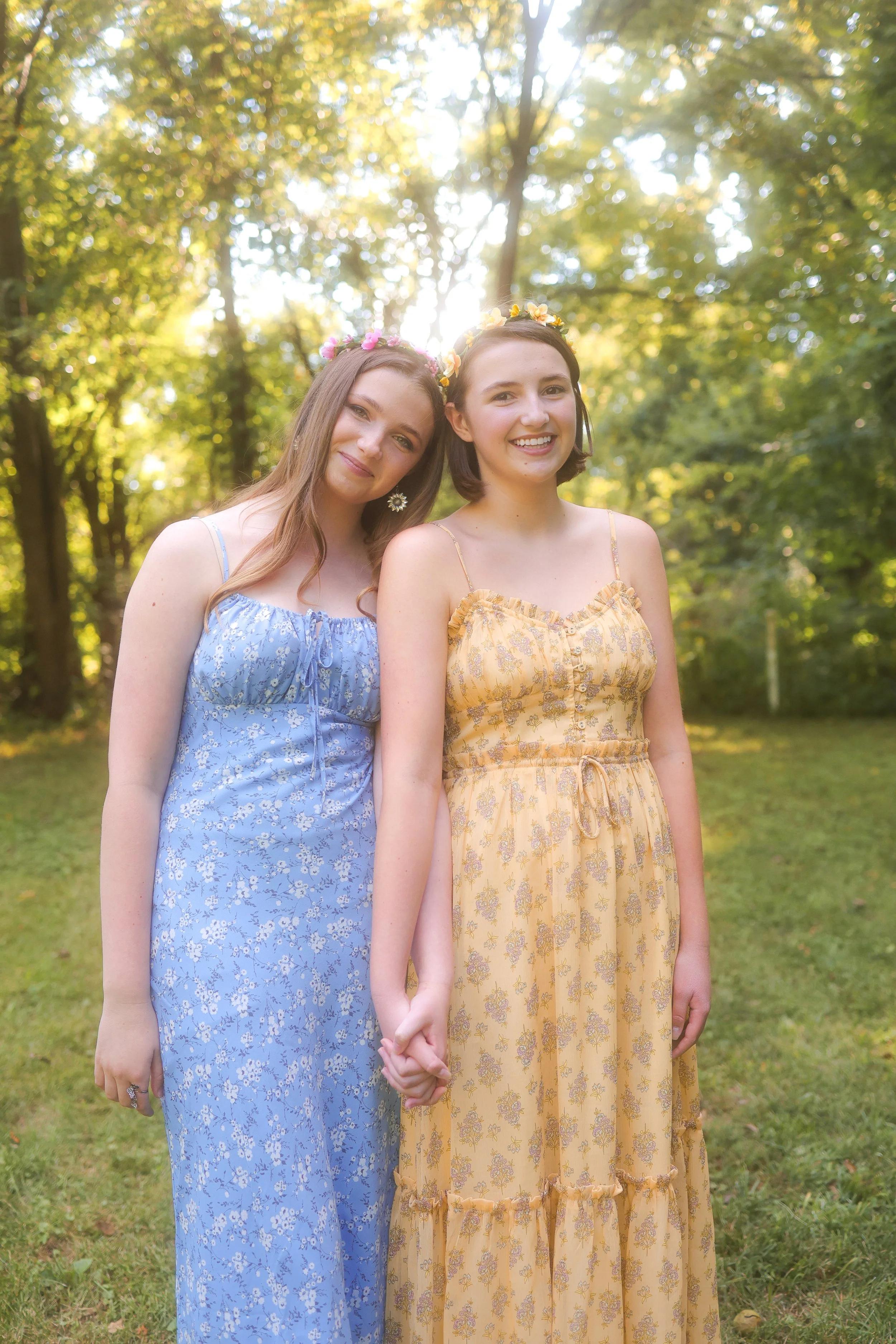 Two young women holding hands and smiling outdoors in a park with sunlight shining through trees during portrait photo session in Carmel, IN.