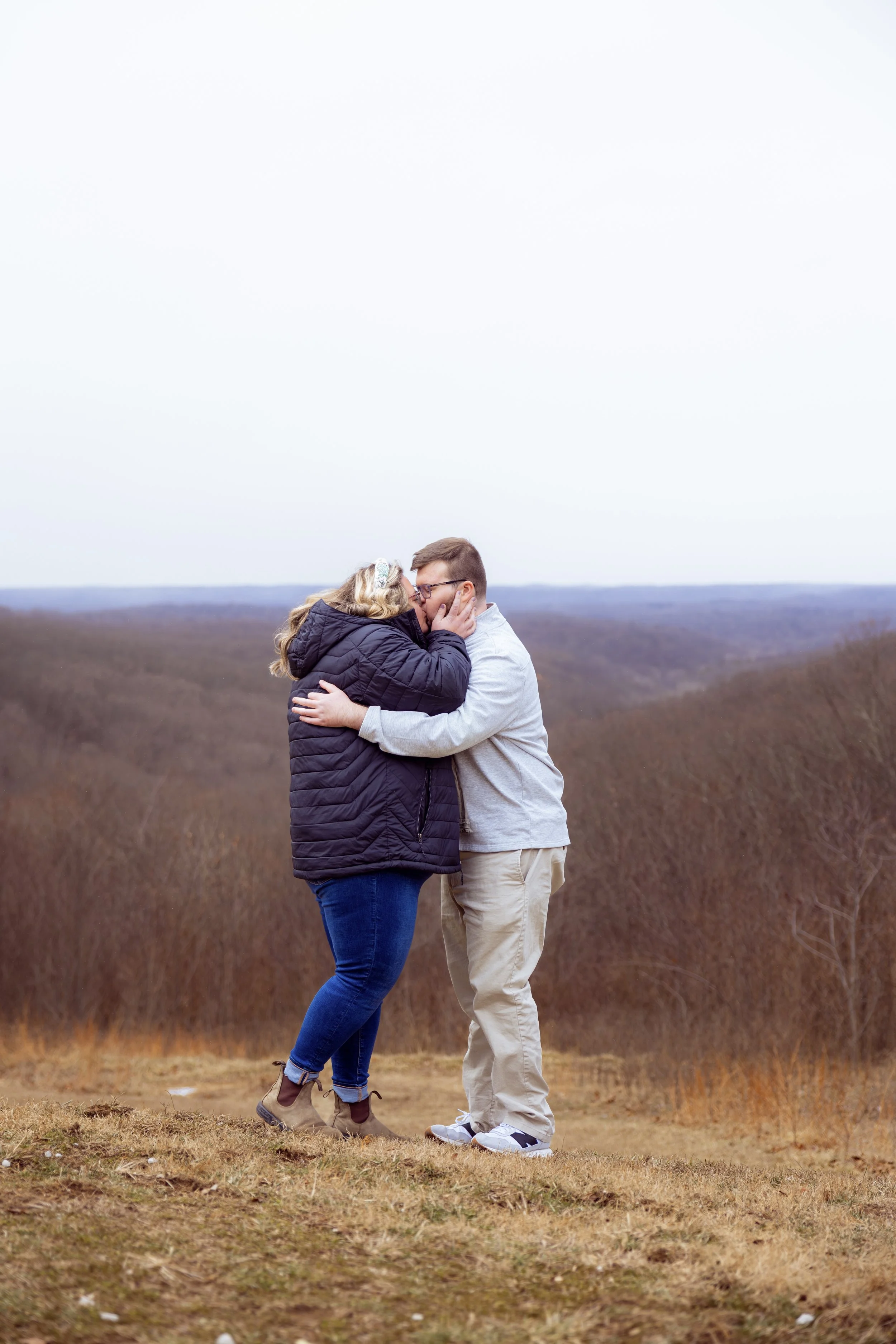Couple sharing a kiss outdoors on a cloudy day with a landscape of trees and hills in the background after the man proposed