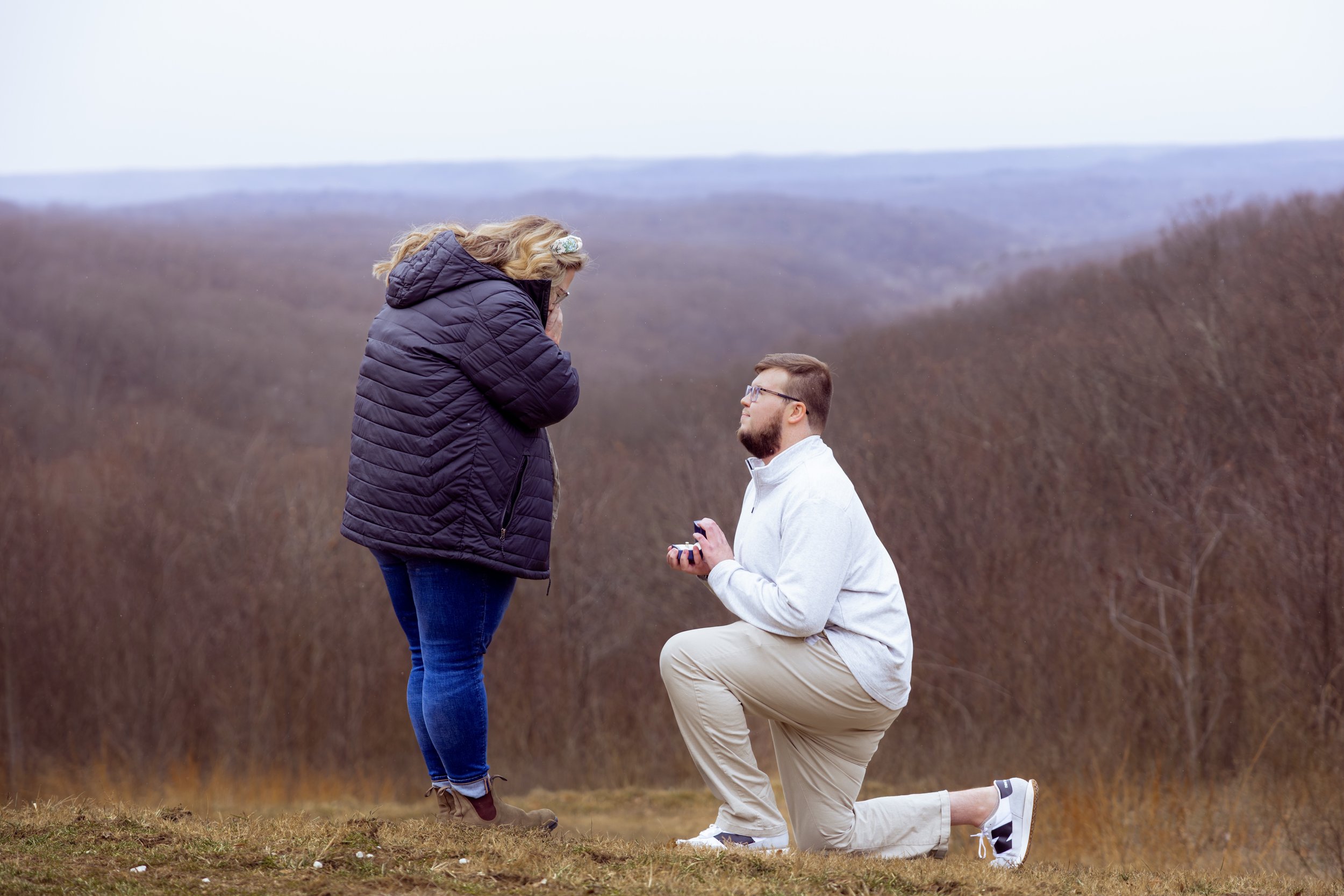 A man proposing marriage to a woman outdoors during daytime, with a scenic view of hills and trees in the background.