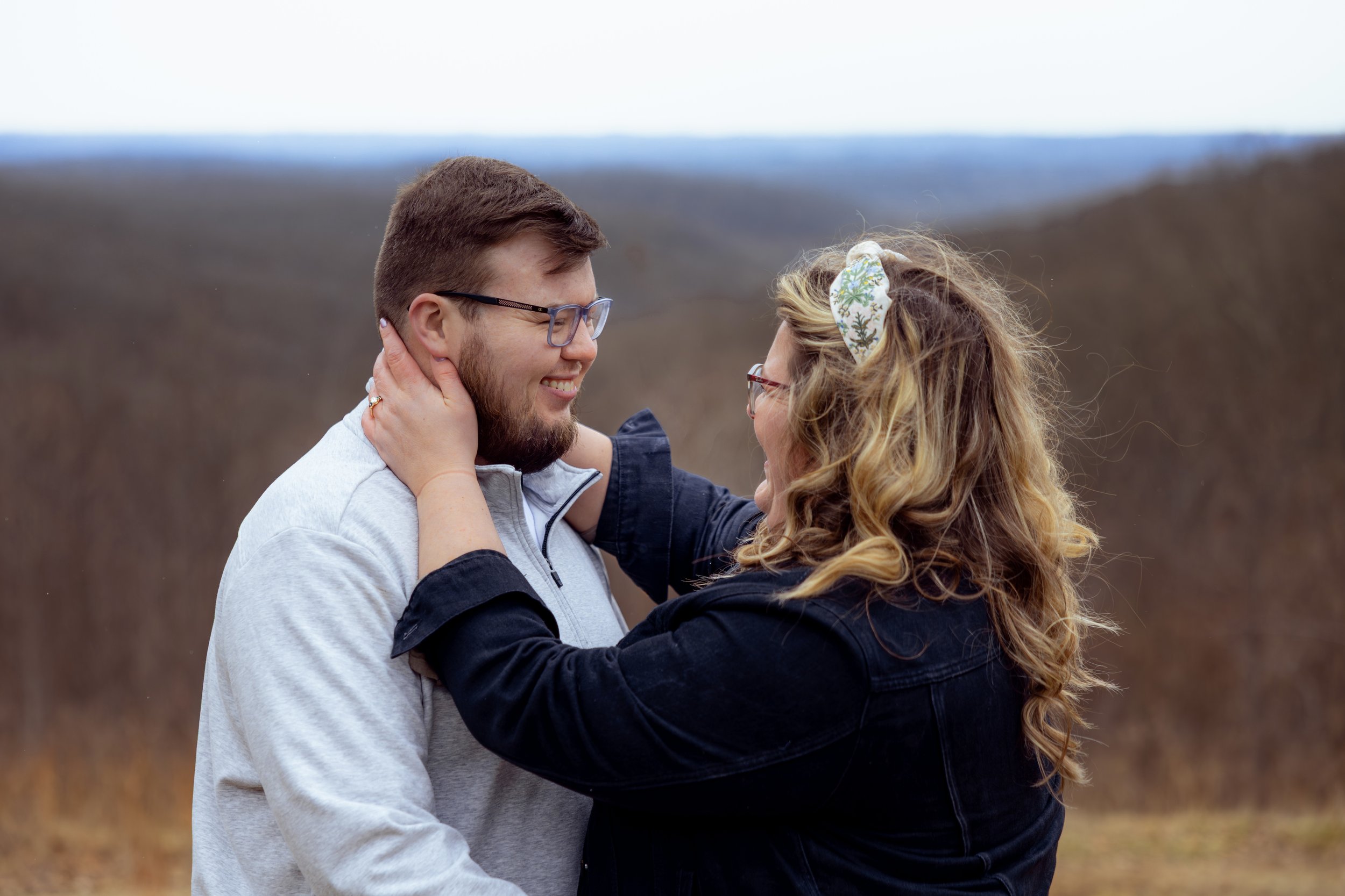 A couple embracing outdoors on a cloudy day, with the woman holding the man's face and smiling at each other.