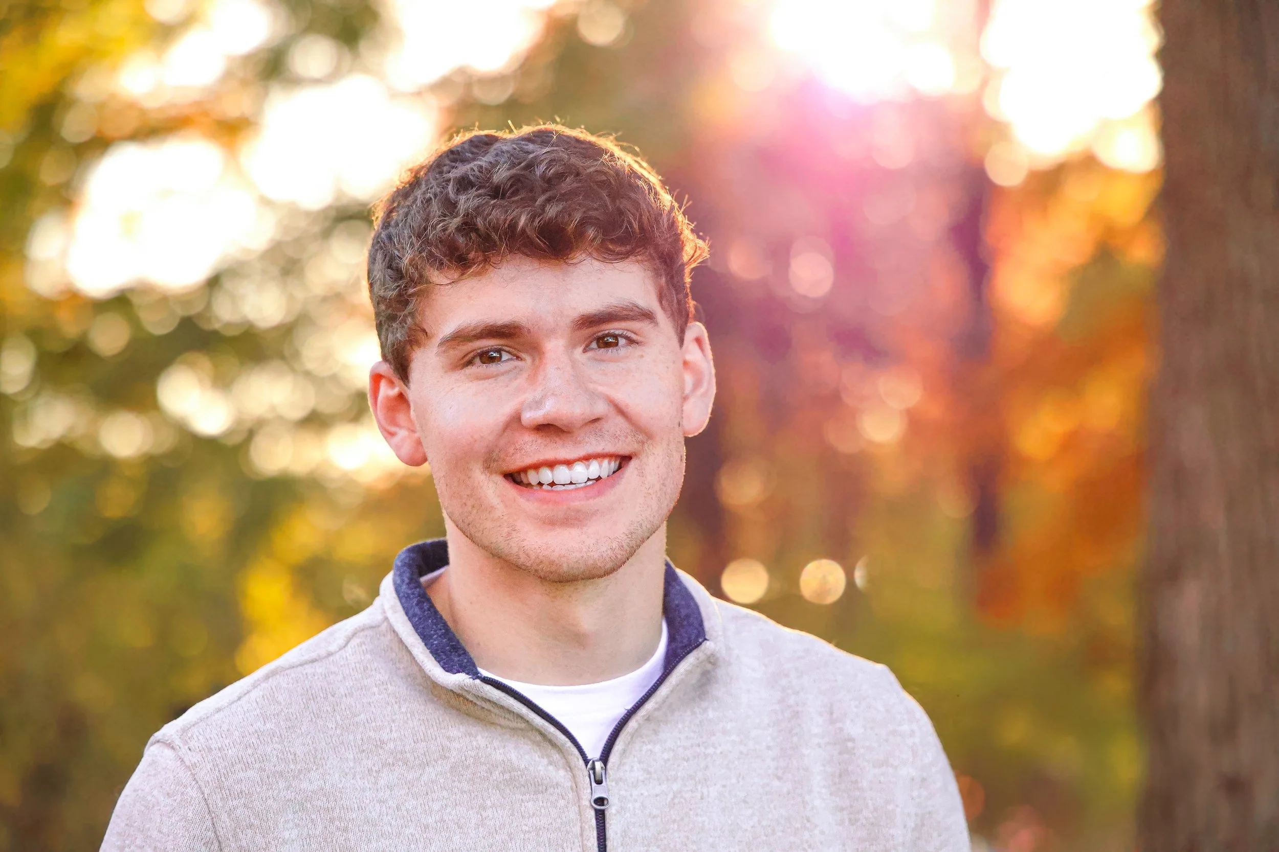 A young man smiling outdoors during autumn, with colorful fall foliage in the background.