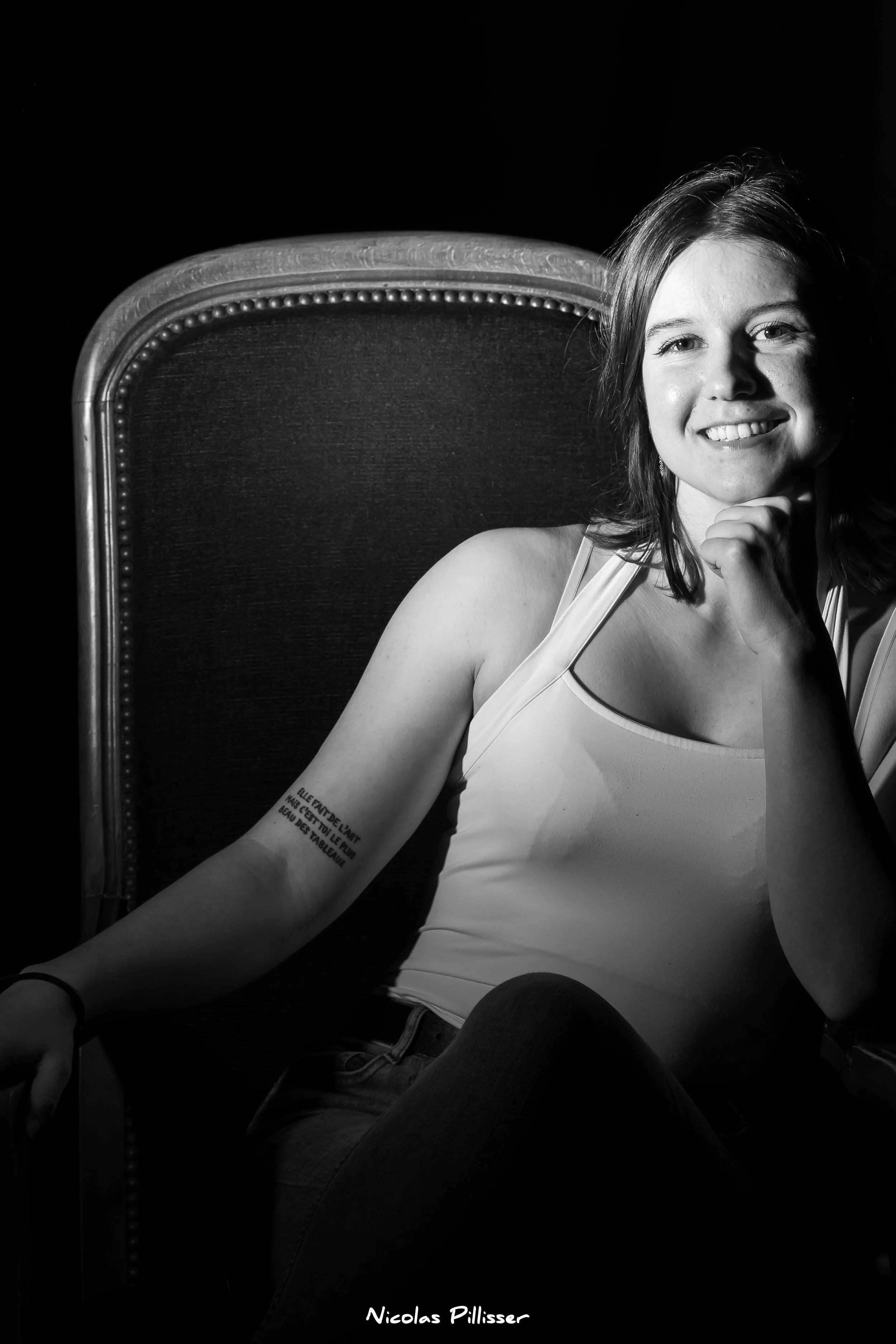 Jeune femme souriante assise sur une chaise, en noir et blanc, avec un tatouage sur le bras gauche. Fond noir, elle porte une haut blanc. Elle a les cheveux bruns, longs et détendus. Sa main repose sous son menton, posée sur son visage.