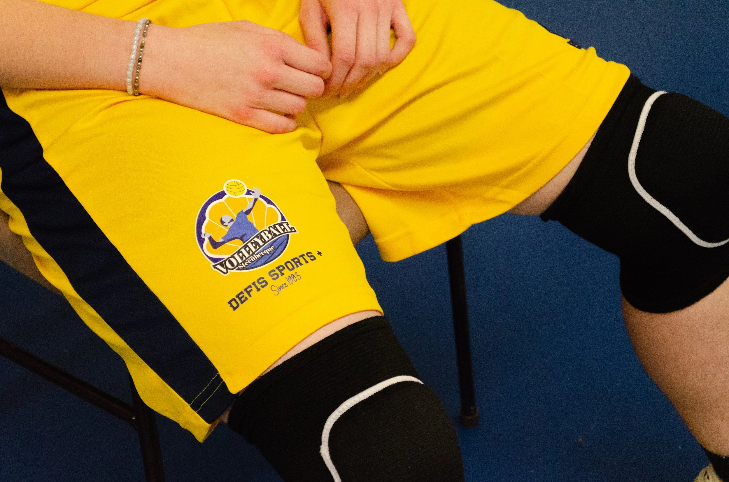 Un joueur de volley-ball portant un maillot jaune avec le logo 'Volleybal' et une décoration en blanc. La personne porte également un bracelet à perles et une chaussette noire avec une bande blanche. Le fond est bleu.