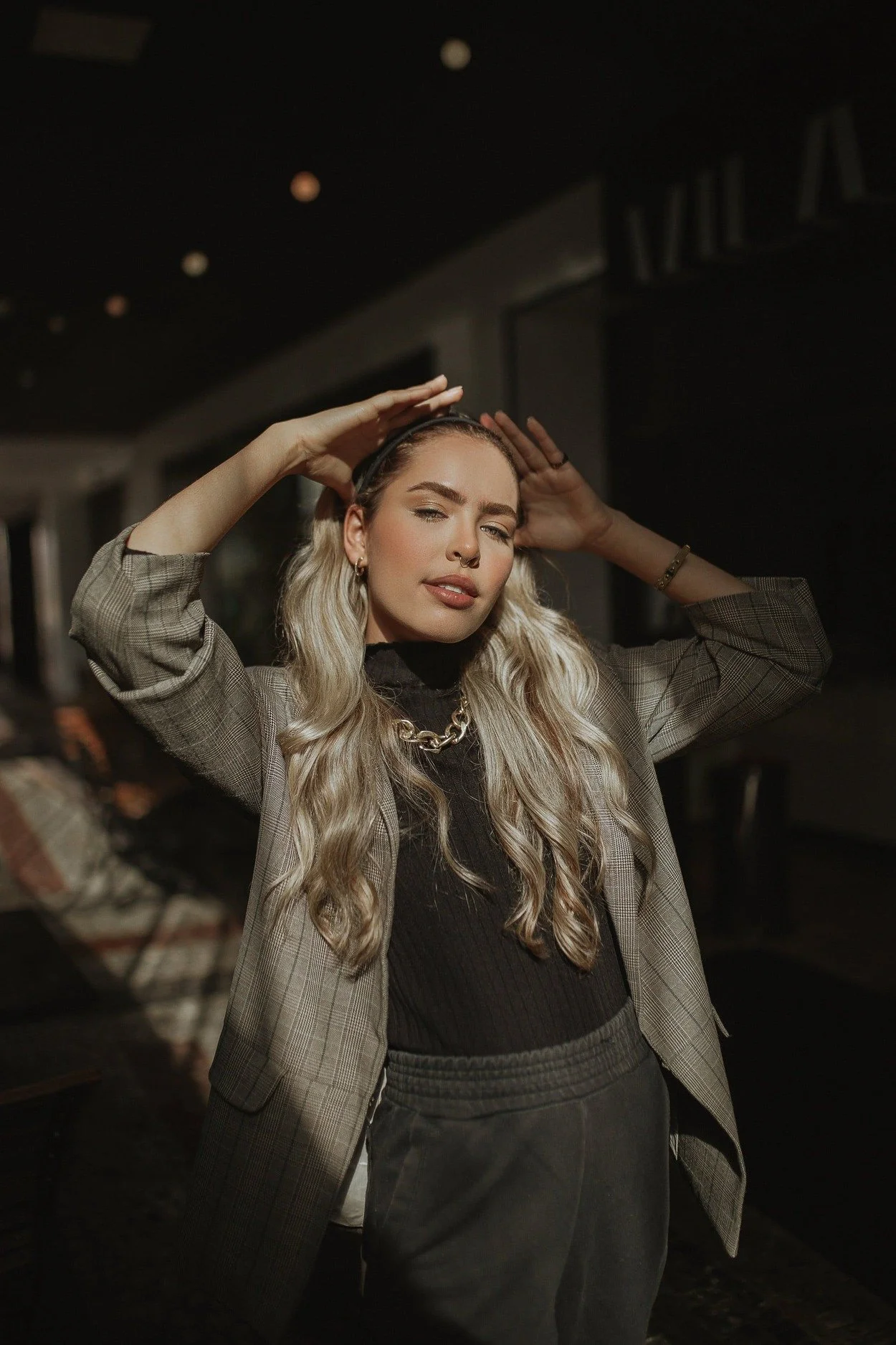 A young woman with long blonde hair standing indoors, holding her hair with both hands, wearing a plaid blazer, black top, and gold chain necklace, in a dimly lit setting.