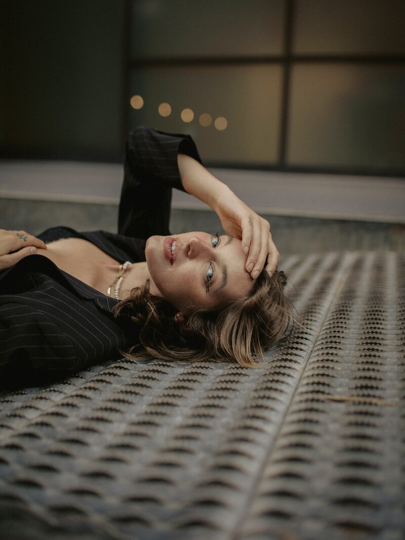 A woman with light brown hair and blue eyes lying on a patterned floor, wearing a black pinstripe blazer, with her hand on her forehead, looking at the camera.