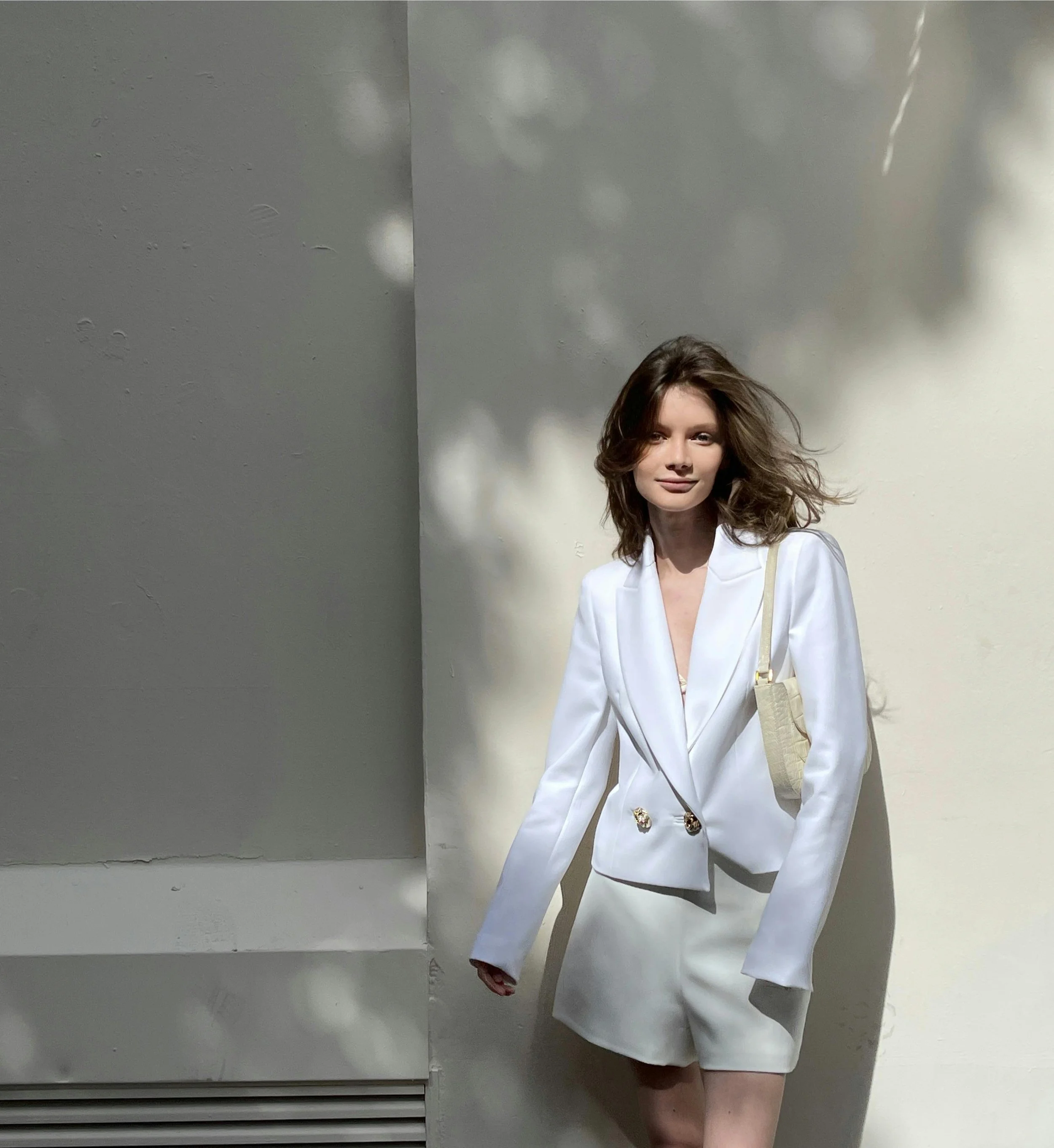 Young woman in a white blazer and shorts standing against a white wall with shadows, carrying a beige shoulder bag.