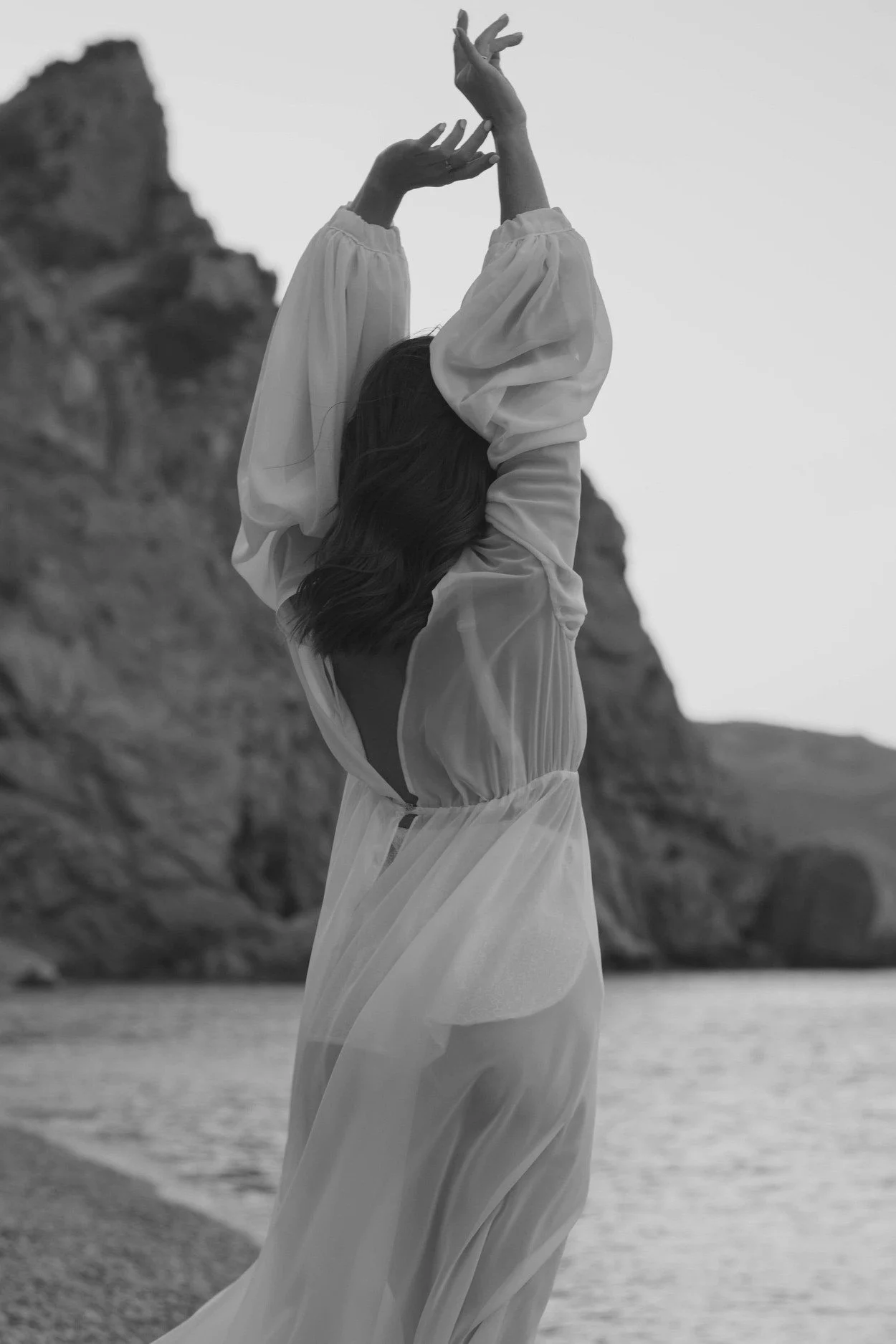 A woman in a flowing sheer dress with her back to the camera, standing near water and rocky cliffs in the distance, with her arms raised above her head.