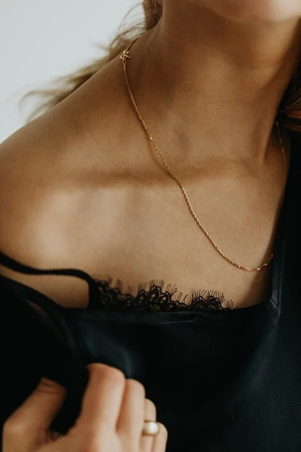 Close-up of a woman's collarbone area, showing a gold chain necklace and black lace clothing. She is holding the fabric with her hand, and part of her face and hair are visible.