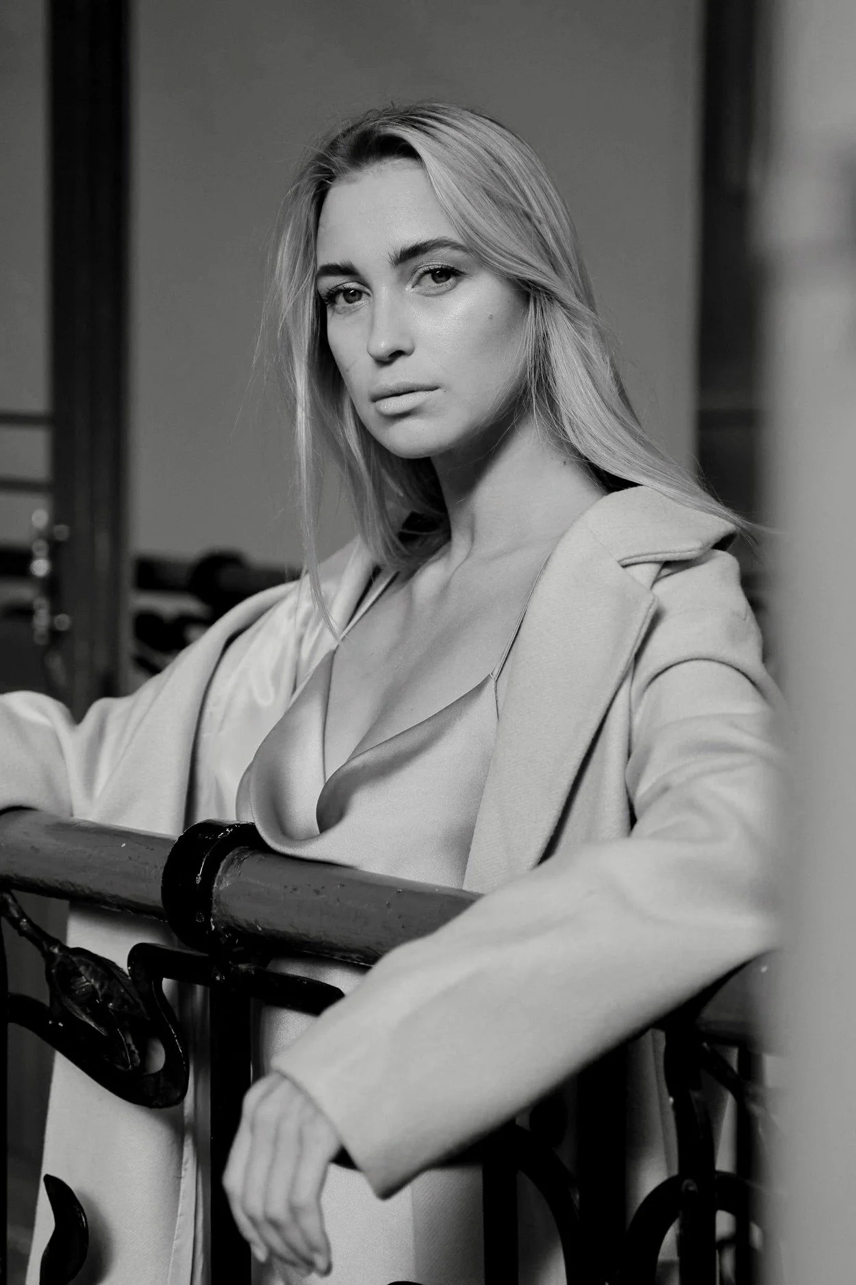 Black and white portrait of a woman with long blonde hair, wearing a blazer and a blouse, standing behind a railing, looking at the camera in a studio setting.