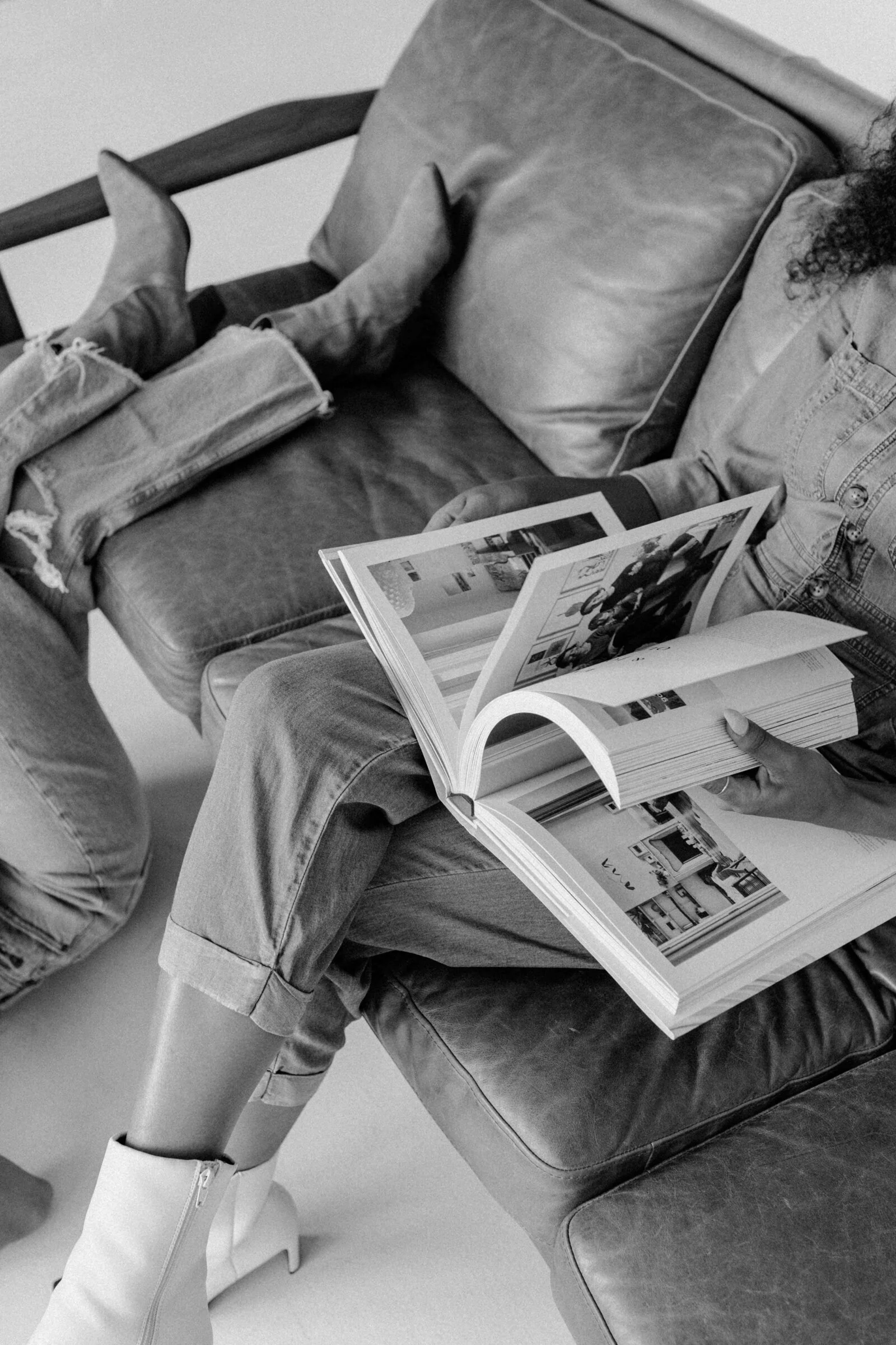Person sitting on a leather couch reading a large photo book, wearing jeans and white boots.