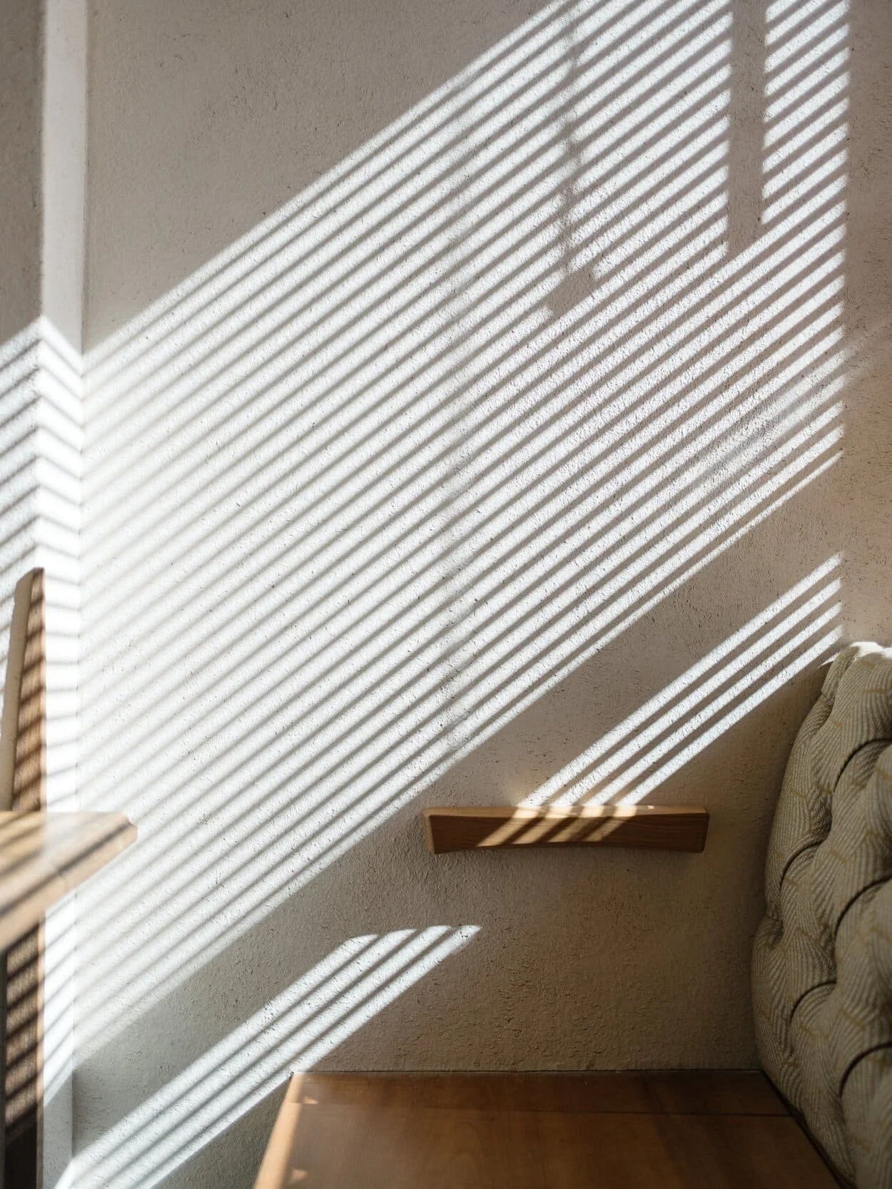 Sunlight streaming through window blinds, casting striped shadows on a textured wall, part of a wooden ledge, a sofa with patterned fabric, and a wooden table.