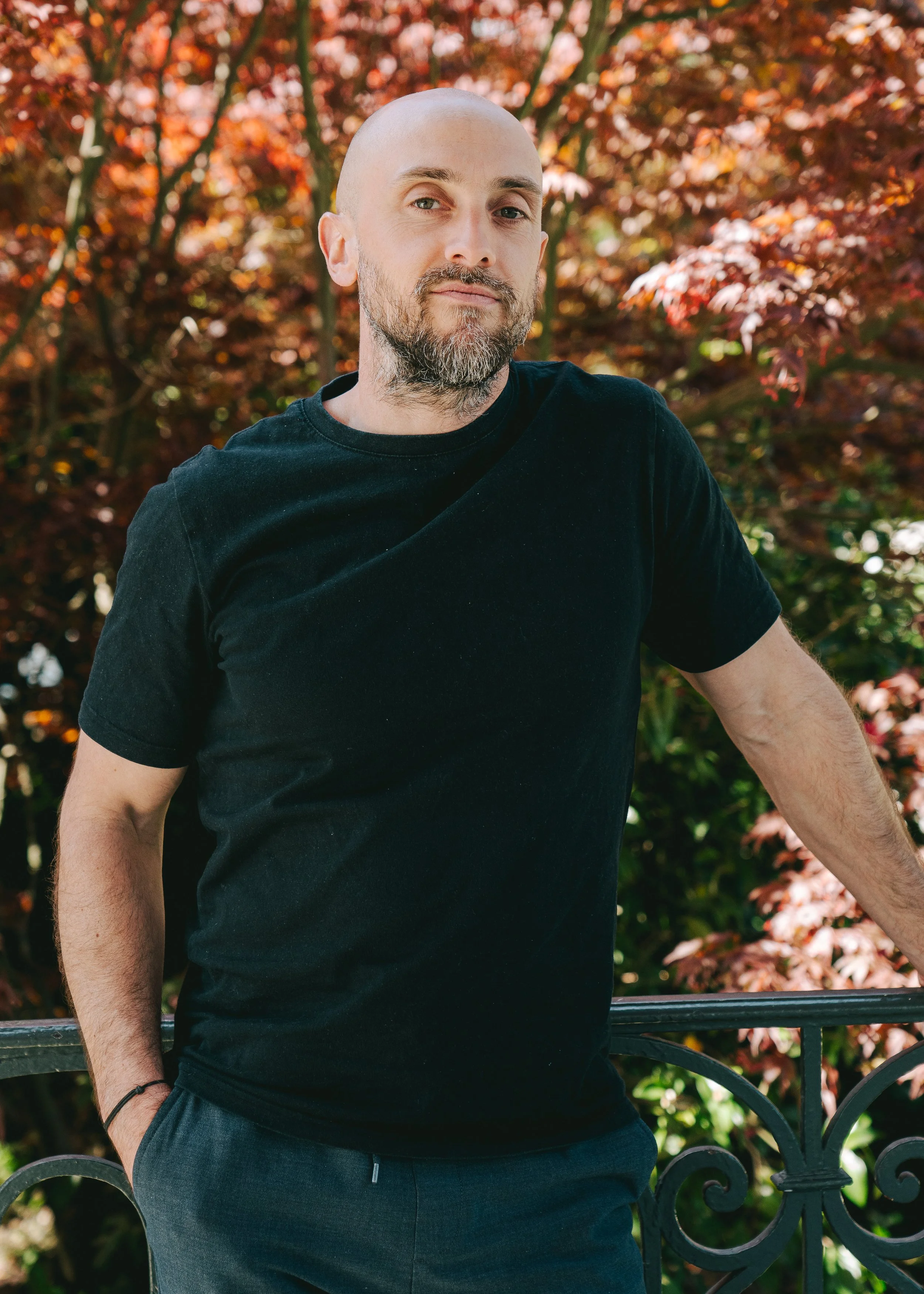 Un homme chauve avec une barbe, portant un t-shirt noir, posant près d'une barrière en fer forgé avec un fond de feuillage d'automne aux couleurs rouges et orange.