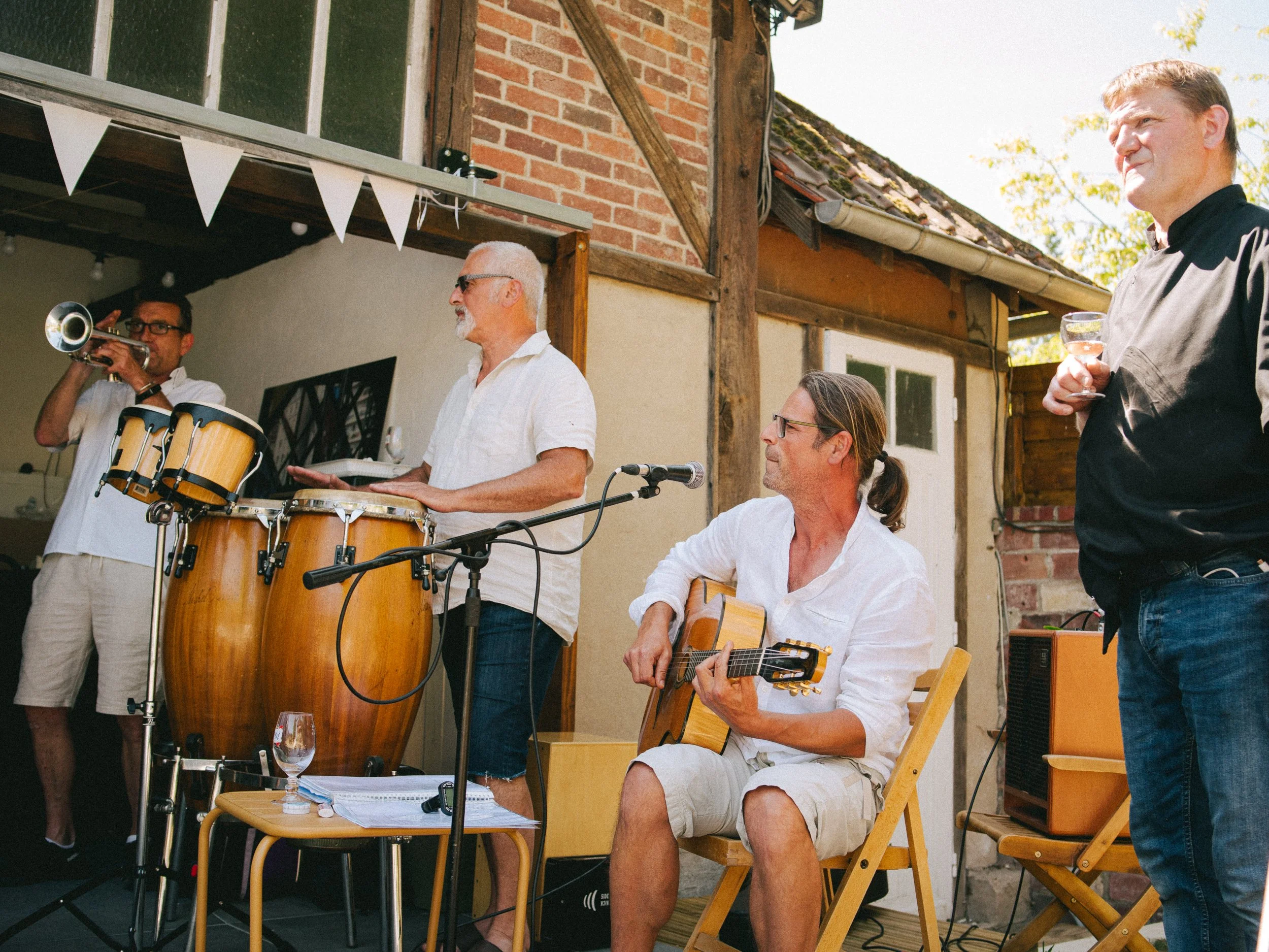 Groupe de musiciens jouant lors d'une fête en extérieur, avec une personne jouant de la guitare, une autre au tambour, un trompettiste et une personne tenant un verre. Une ambiance en plein air avec un bâtiment en arrière-plan.