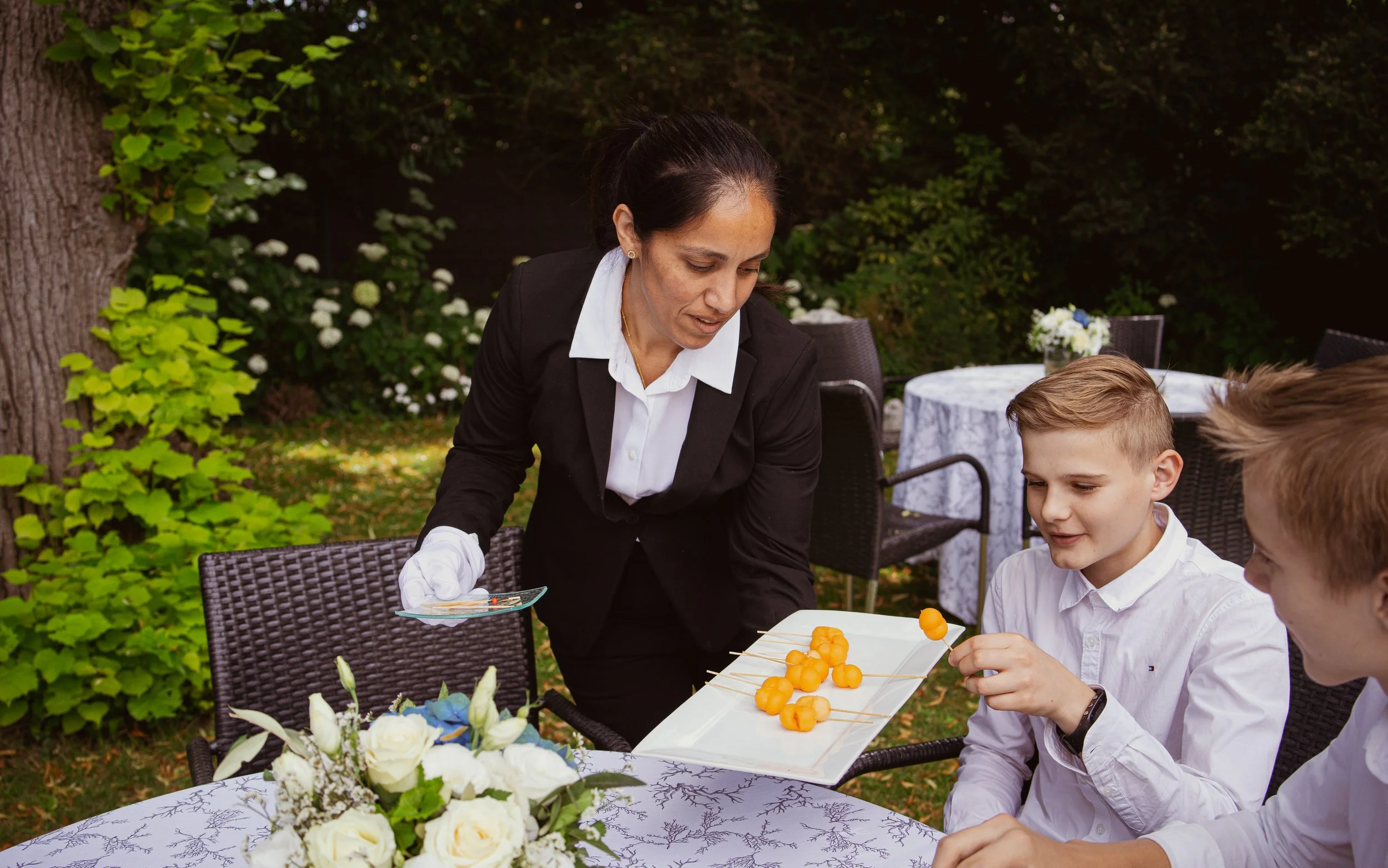 Une femme serveuse utilisant des gants blancs sert des amuse-gueules sur des piques à une table lors d'un événement en plein air, avec deux jeunes hommes habillés en blanc. Il y a des fleurs blanches sur la table, entourée d'arbres et de chaises.