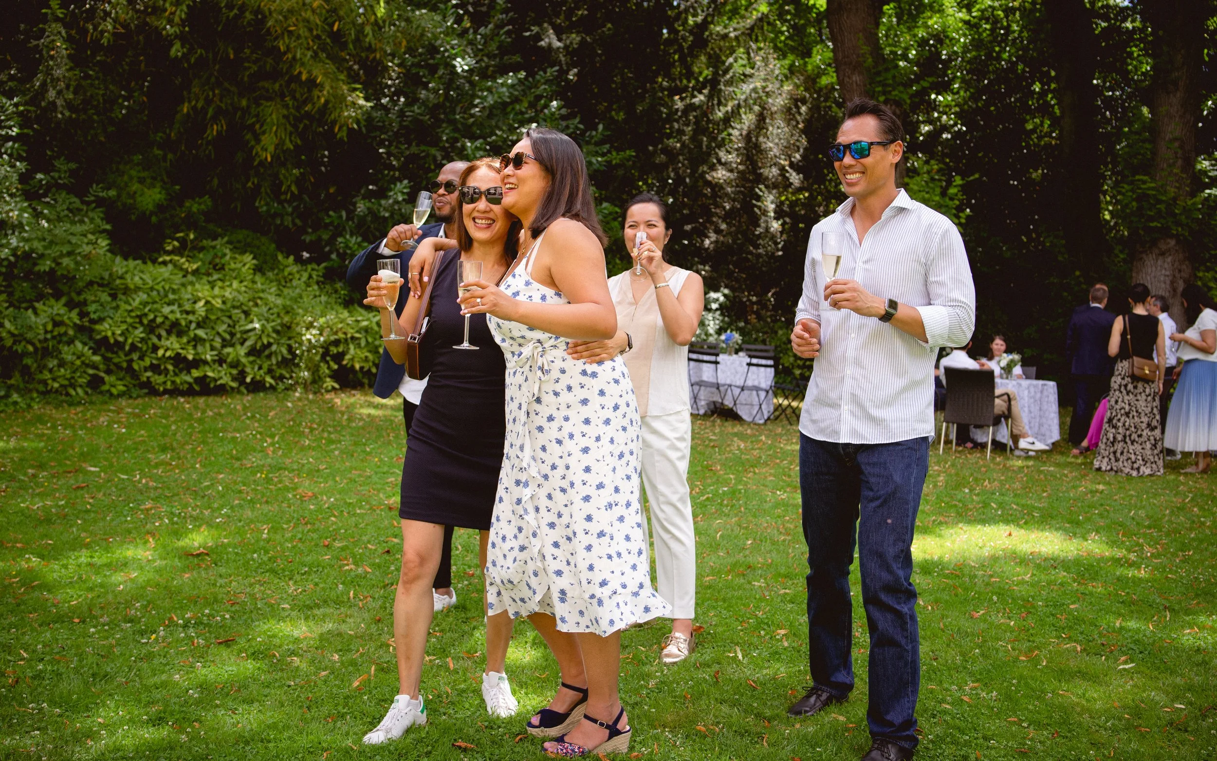 Groupe de personnes souriantes célébrant lors d'une fête en plein air, tenant des flûtes de champagne sur une pelouse verte avec des tables et des invités en fond, sous un ciel ensoleillé.