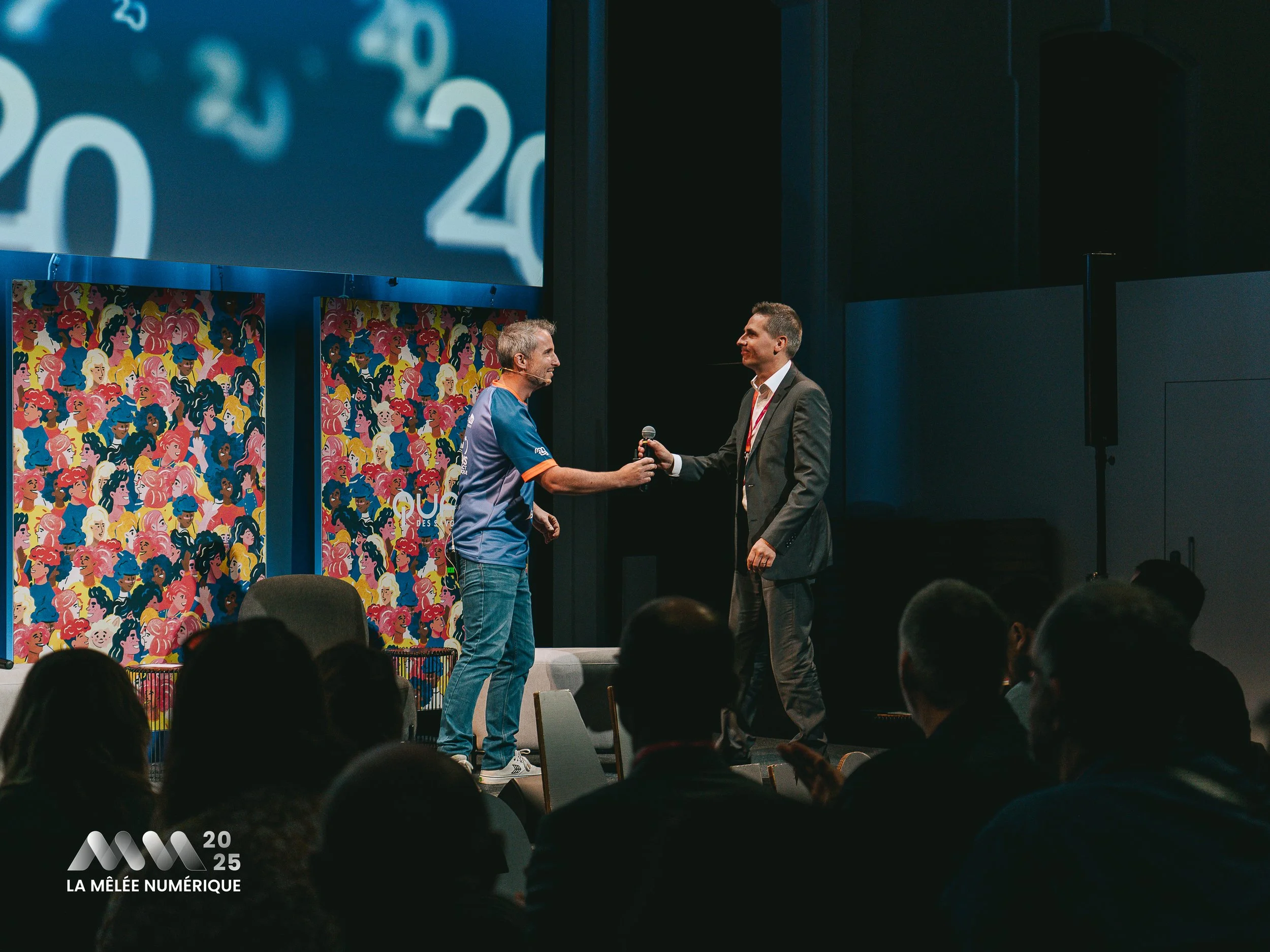 Un homme en uniforme de sport sur scène reçoit un micro d'un homme en costume lors d'une conférence ou événement en 2025, avec un public en arrière-plan et des affiches colorées en fond.