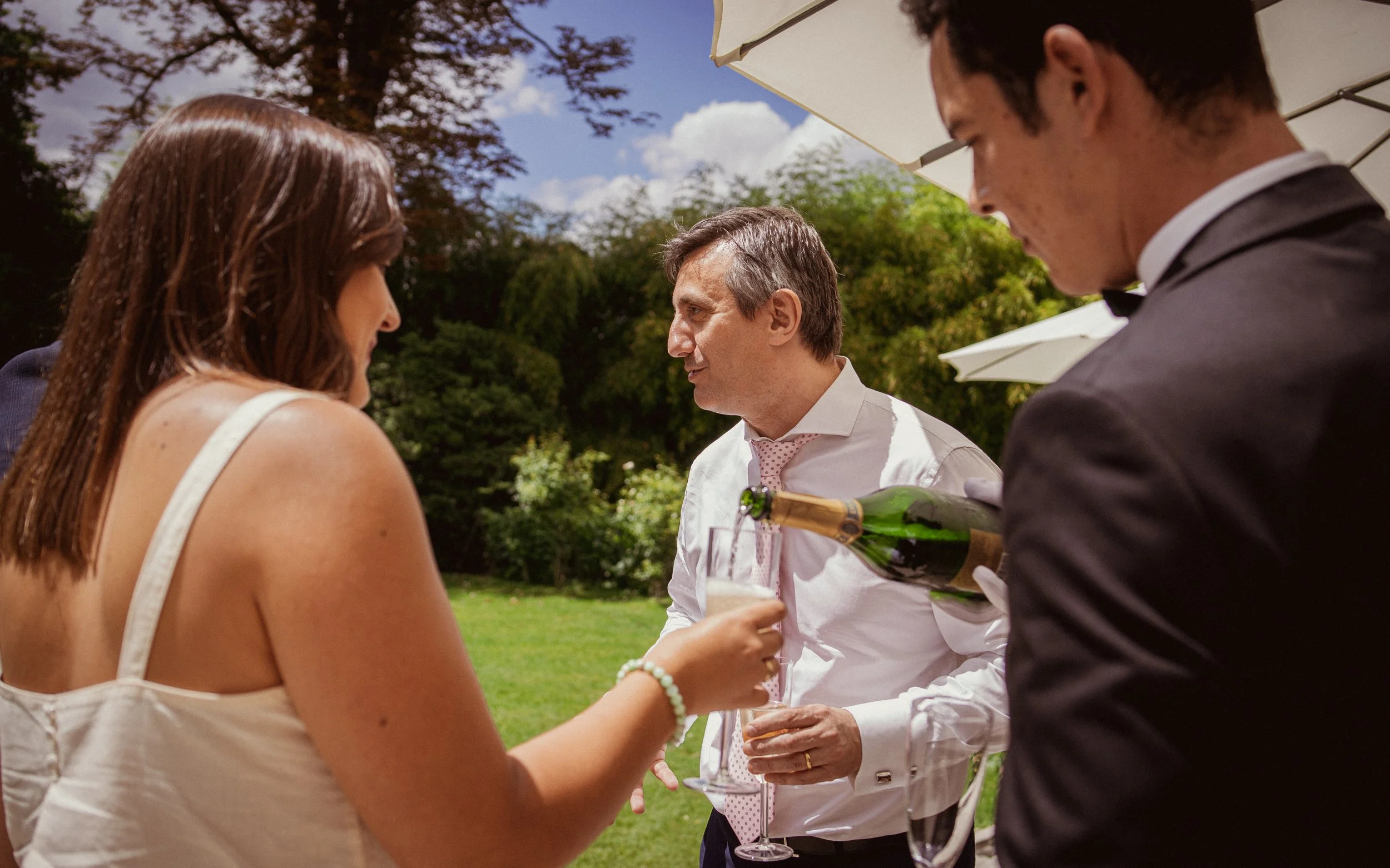 Trois personnes lors d'une fête en plein air, deux hommes en costume et une femme, en train de servir et de recevoir du champagne dans un jardin verdoyant, sous un parasol.