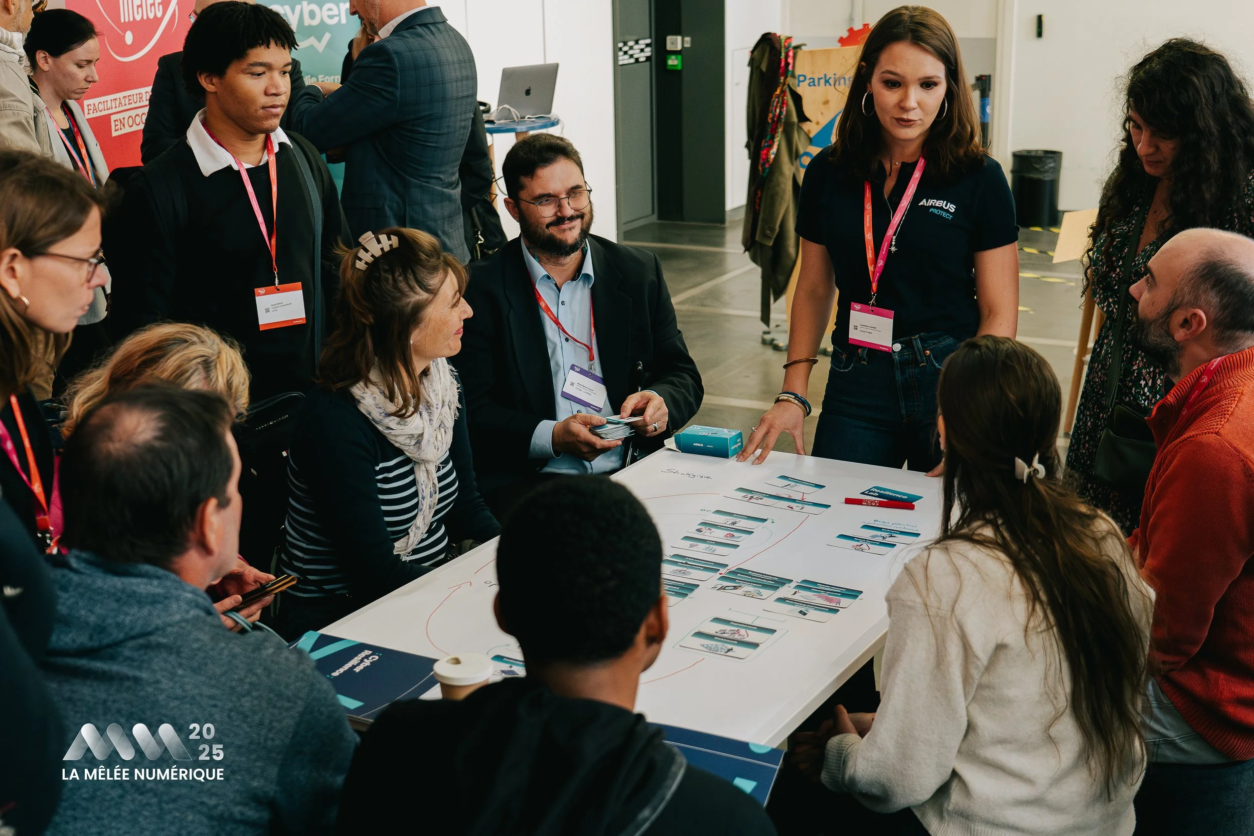 Groupe de personnes assises et debout autour d'une table de conférence lors d'un événement professionnel, en discussion, avec une personne expliquant quelque chose, avec affiches et badges autour.
