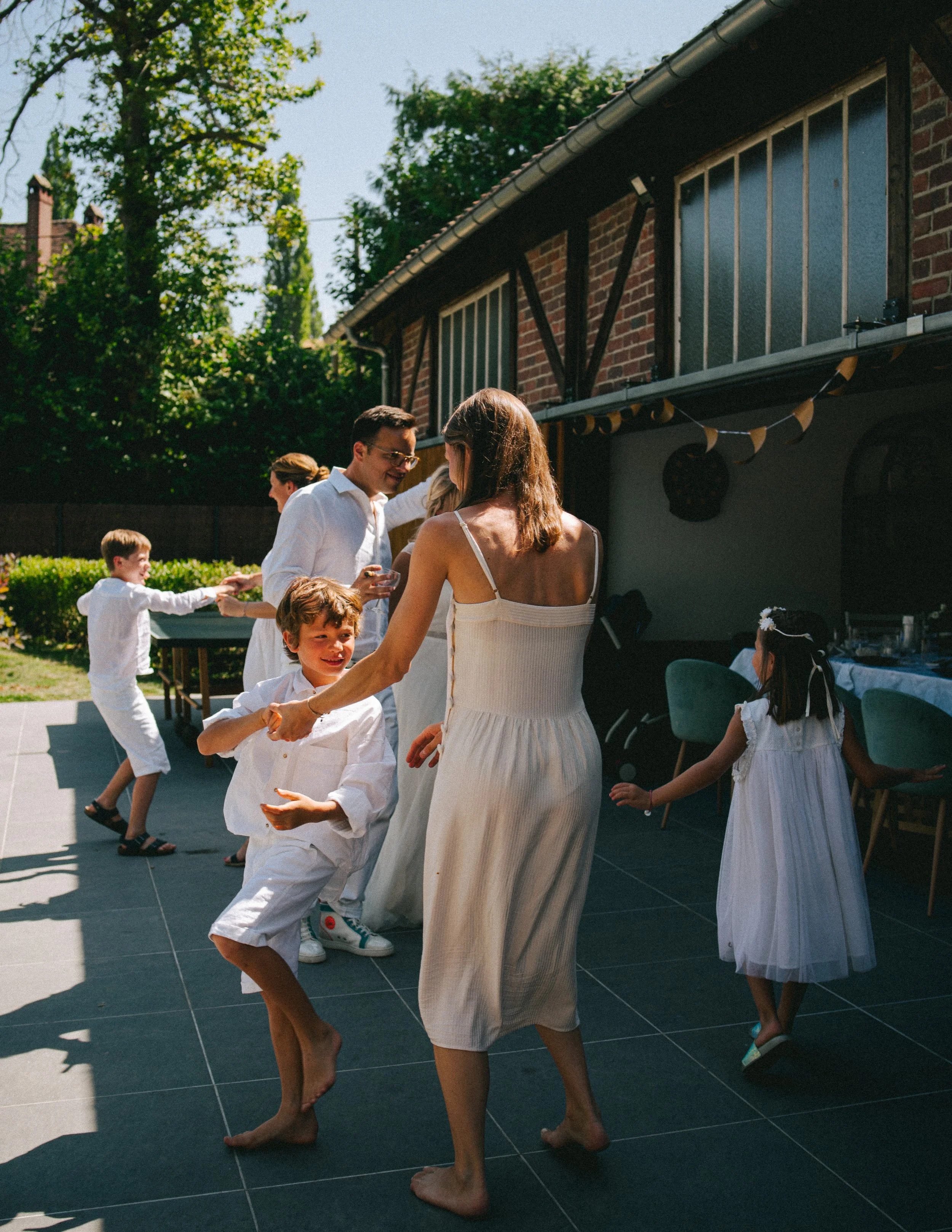 Groupe de personnes, adultes et enfants, dansant lors d'une fête en plein air sur une terrasse ensoleillée, avec une maison en arrière-plan.