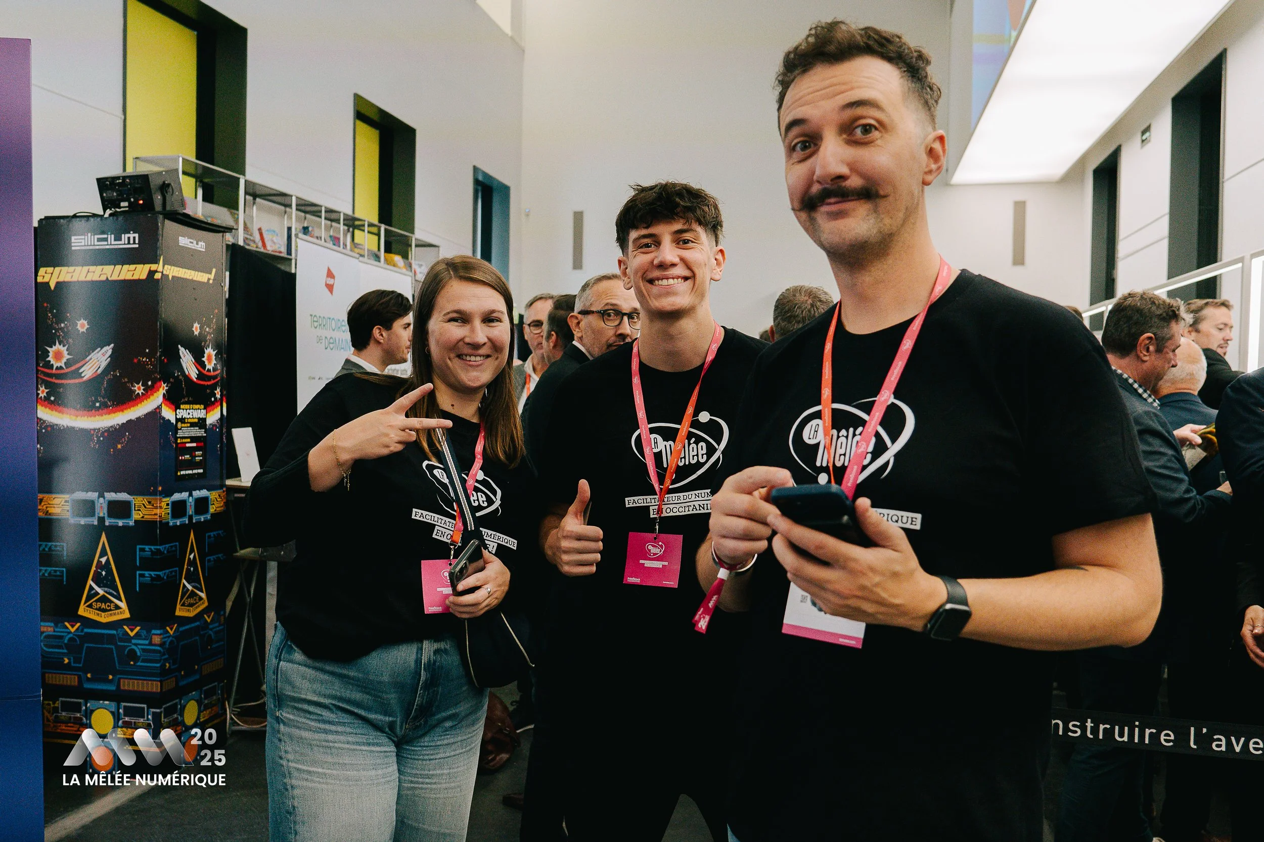 Trois jeunes adultes souriants posent pour une photo lors d'un événement, portant des t-shirts noirs avec un logo et des badges. En arrière-plan, d'autres personnes sont assises et discutent dans une salle lumineuse.