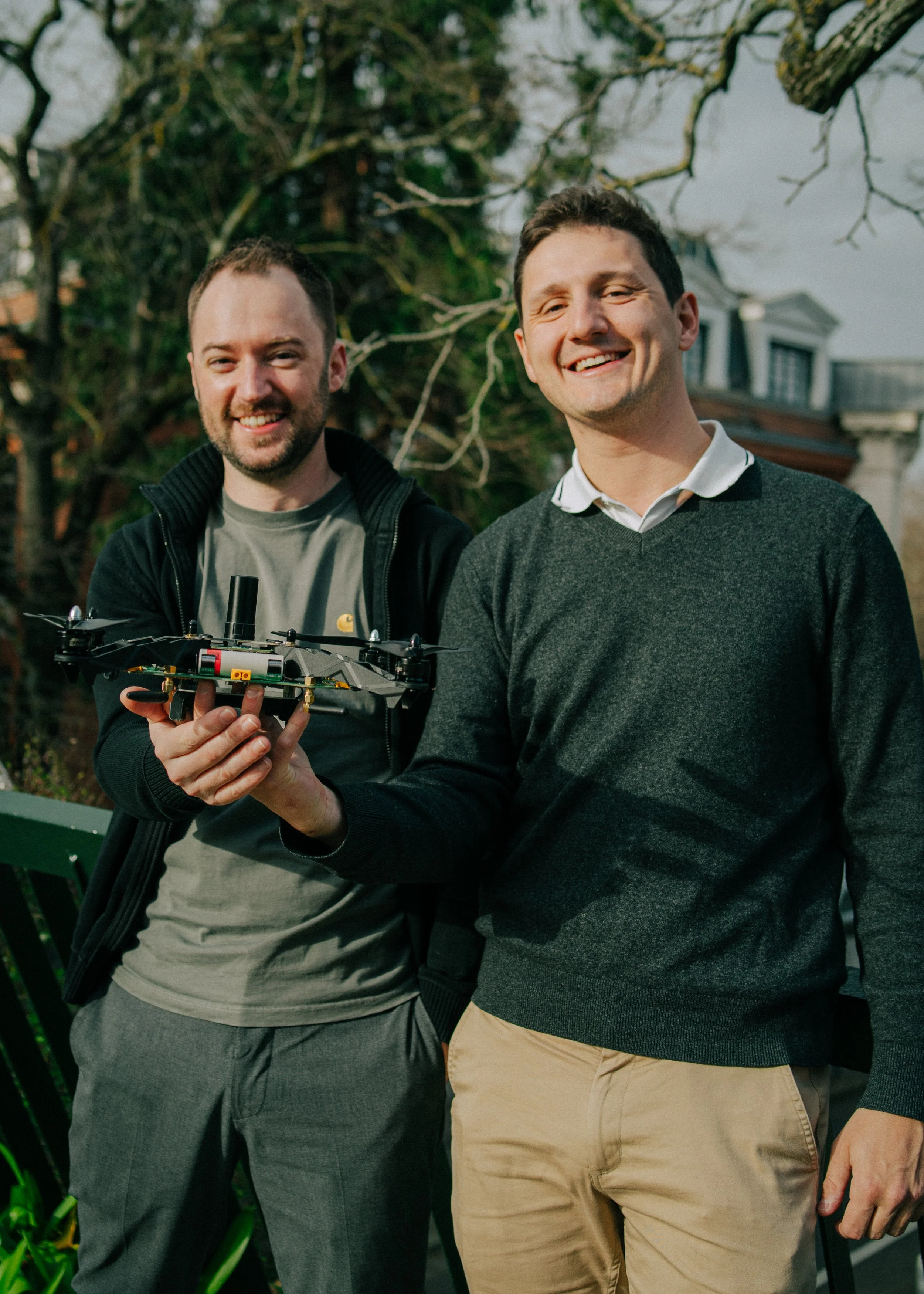 Deux hommes souriants, l'un tenant un drone, dehors, arbres et maisons en arrière-plan.