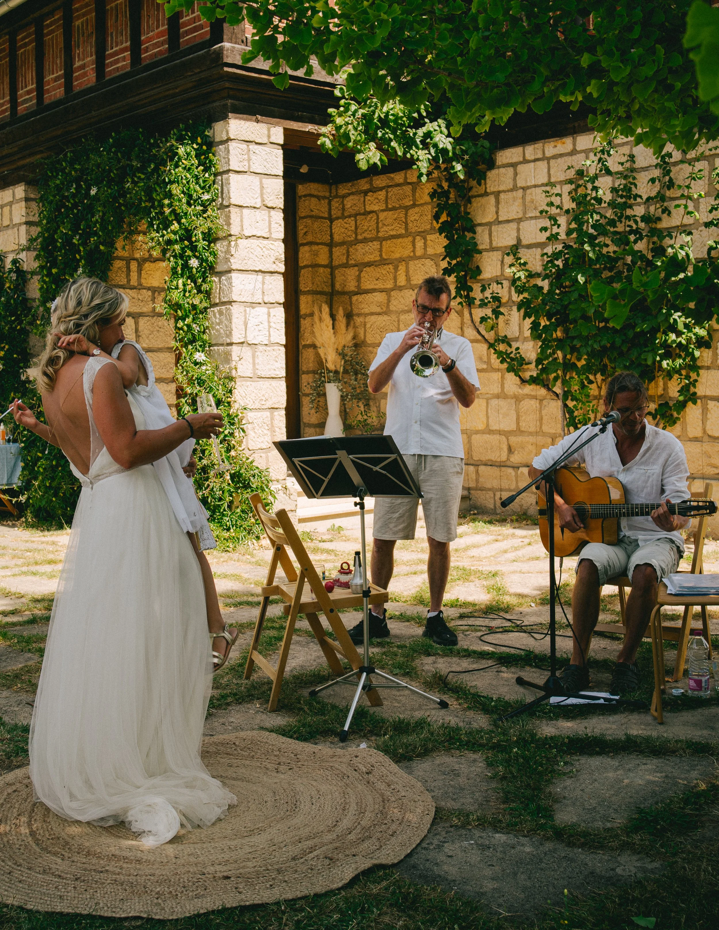 Un groupe de musiciens jouant dans un jardin, avec une femme en robe blanche. Il y a un homme jouant de la guitare, un autre jouant de la trompette, et une femme blanche vêtue d'une robe longue