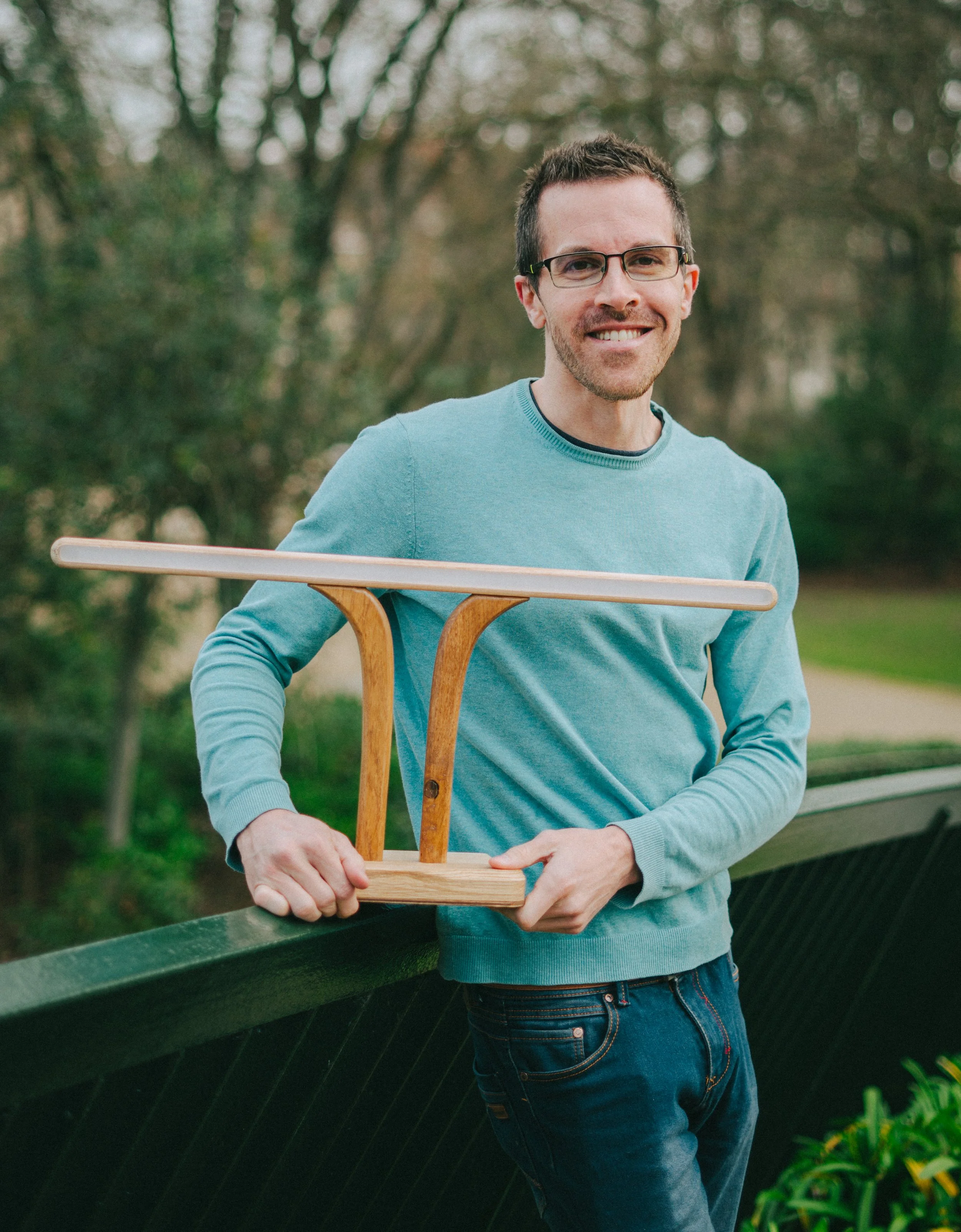 Un homme souriant portant des lunettes, t-shirt bleu, tenant une petite table en bois en équilibre sur une barrière dans un parc.