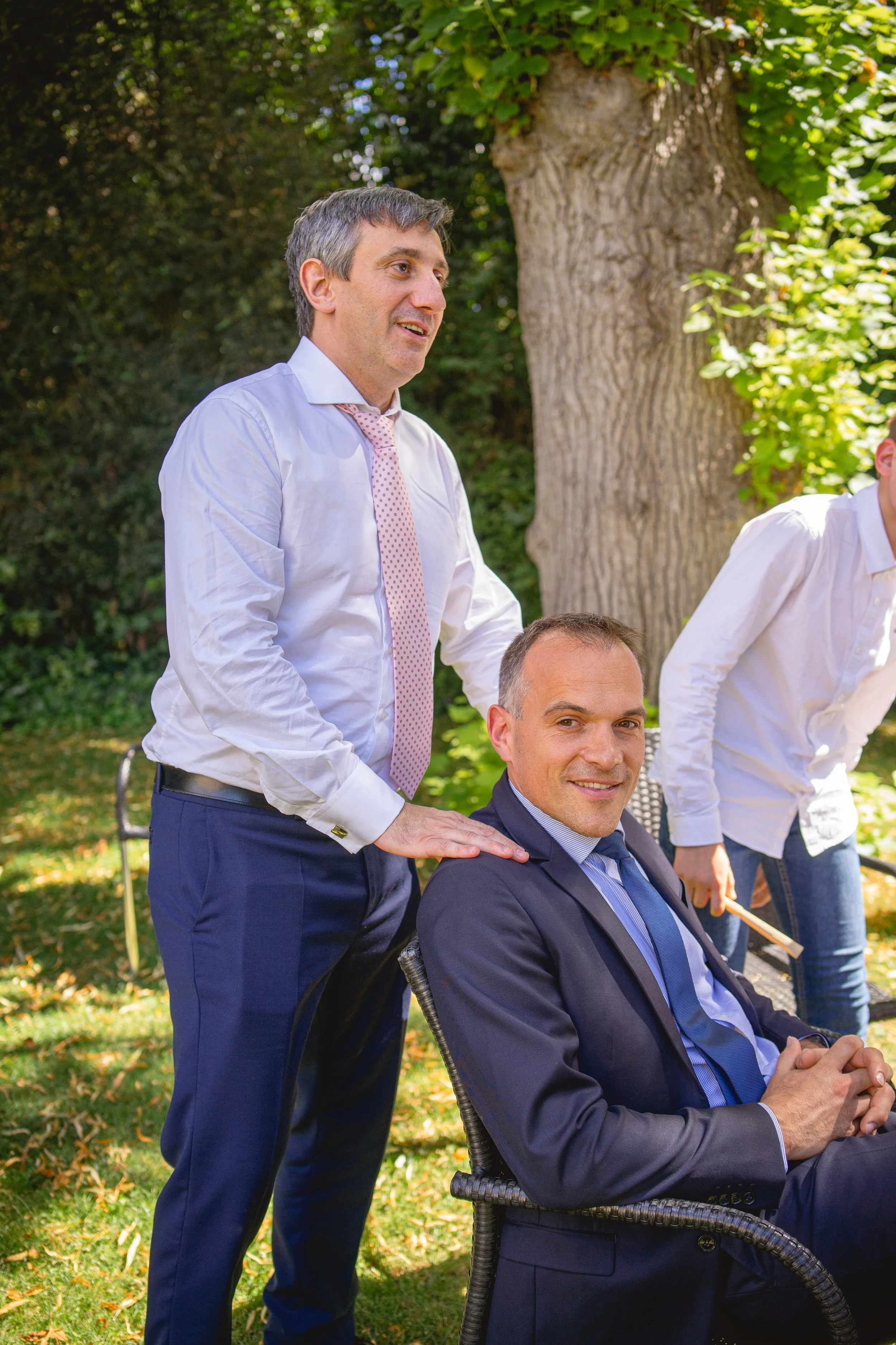 Deux hommes en costume assis et un autre qui leur donne un massage dans un parc ensoleillé.
