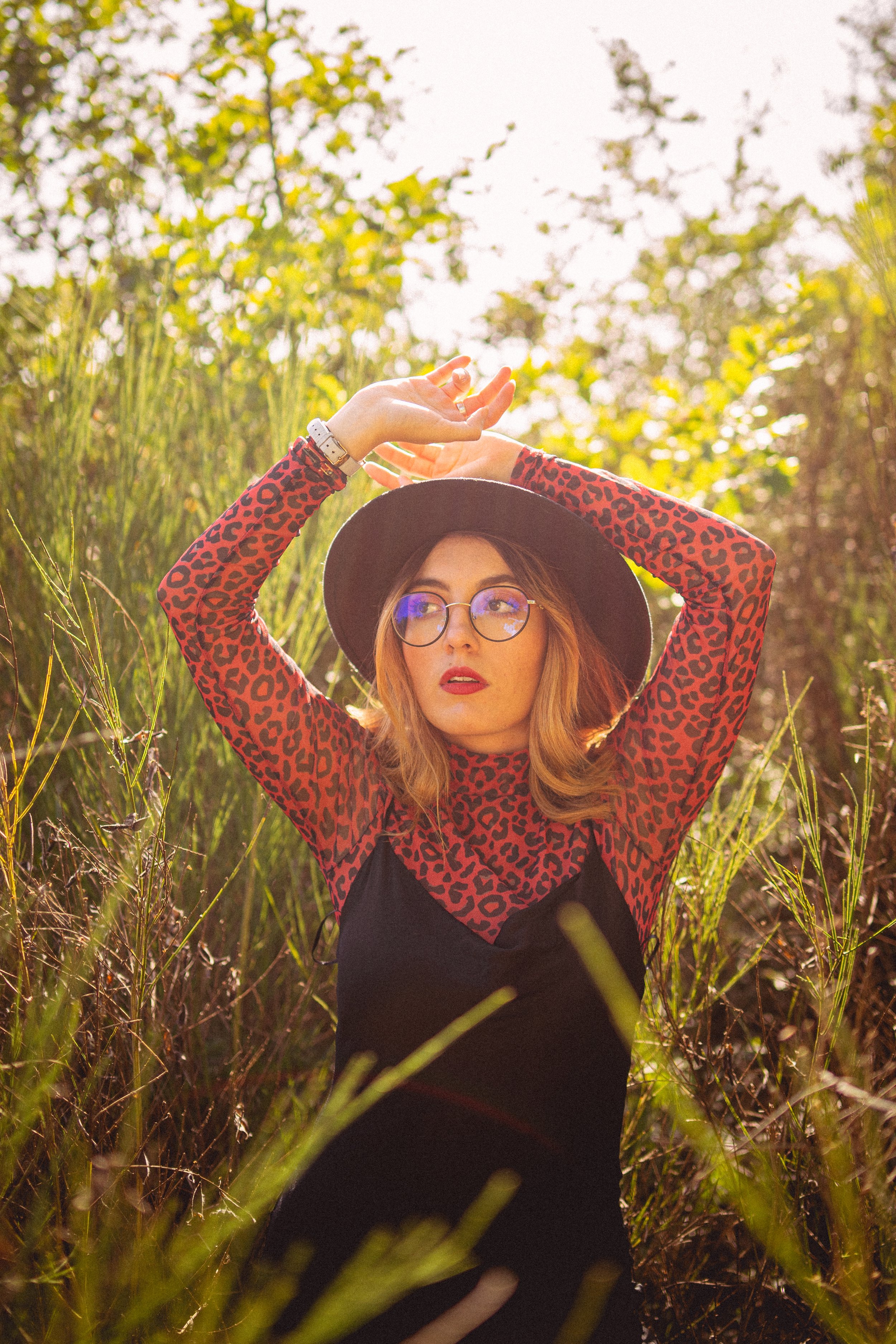 Jeune femme portant un chapeau noir, lunettes rondes, vêtue d'un haut à manches longues à motif léopard rouge et noir, posant dans un champ ensoleillé avec des herbes vertes et des arbres en arrière-plan.
