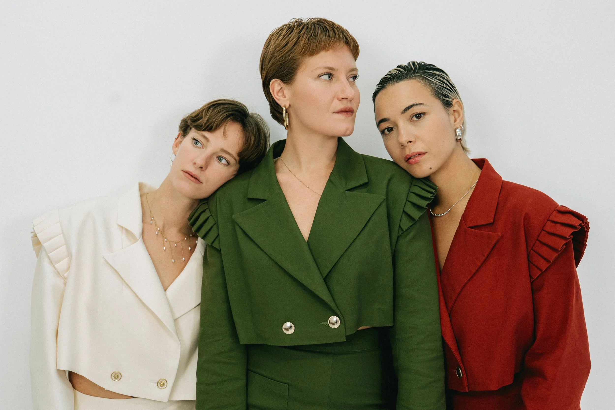 Trois femmes posant contre un mur blanc, portant des vestes colorées avec des détails plissés.