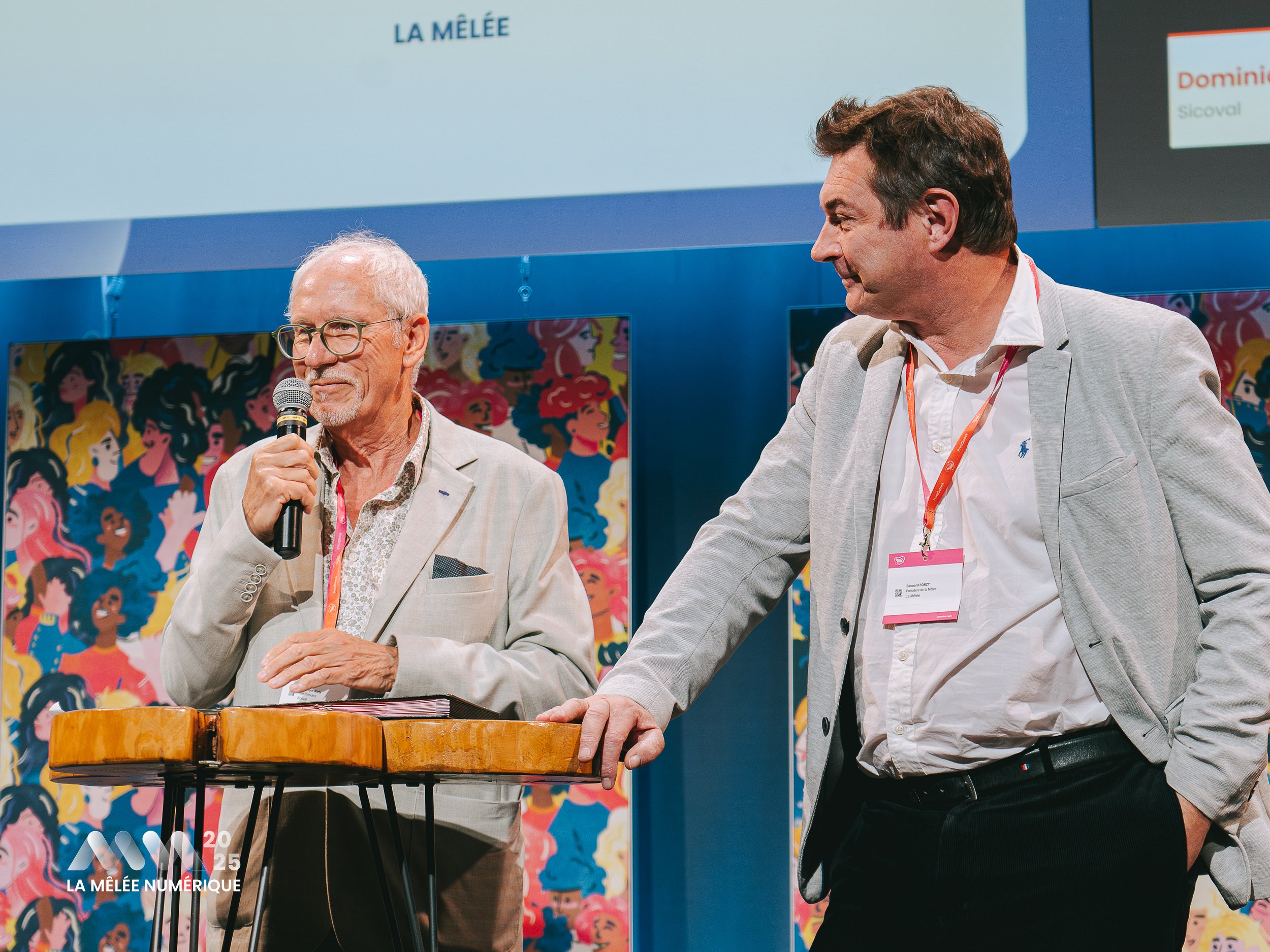 Deux hommes debout sur scène lors d'une conférence : l'un parle en tenant un microphone, l'autre écoute, avec un panneau de fond coloré et un écran affichant 'LA MÉLÉE' en français.