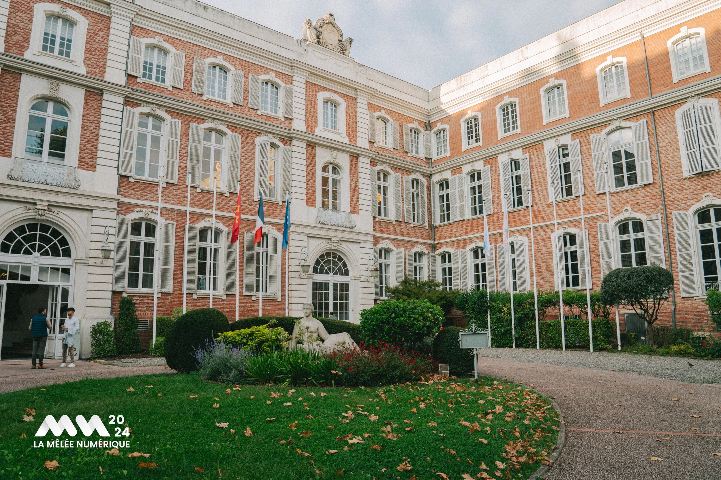Bâtiment historique en briques avec des fenêtres à volets blancs, drapeaux devant, statues et jardin avec arbustes et fleurs.