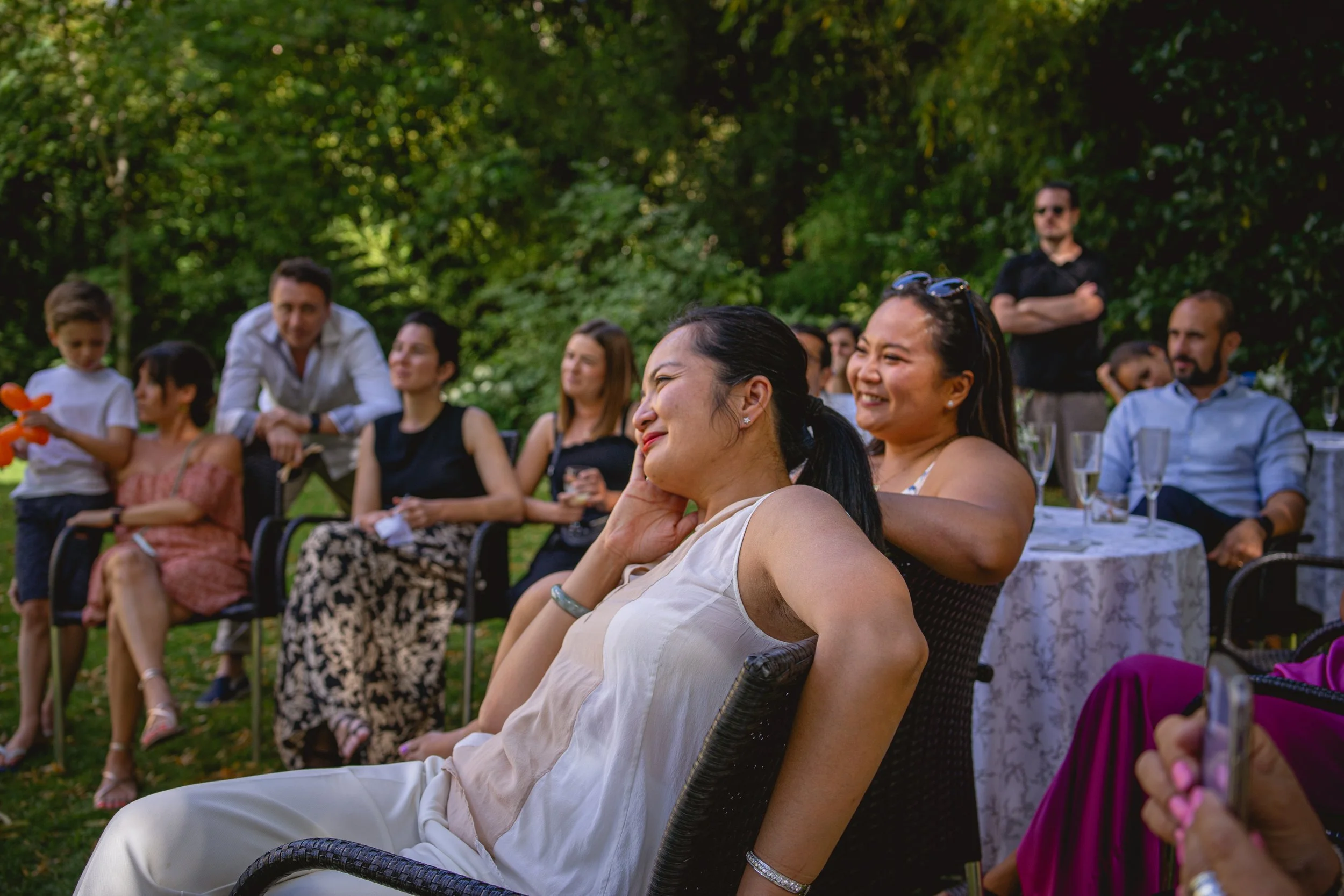 Groupe de personnes souriantes lors d'une réunion ou célébration en plein air, assises autour de tables et sur des chaises dans un jardin avec des arbres en arrière-plan.