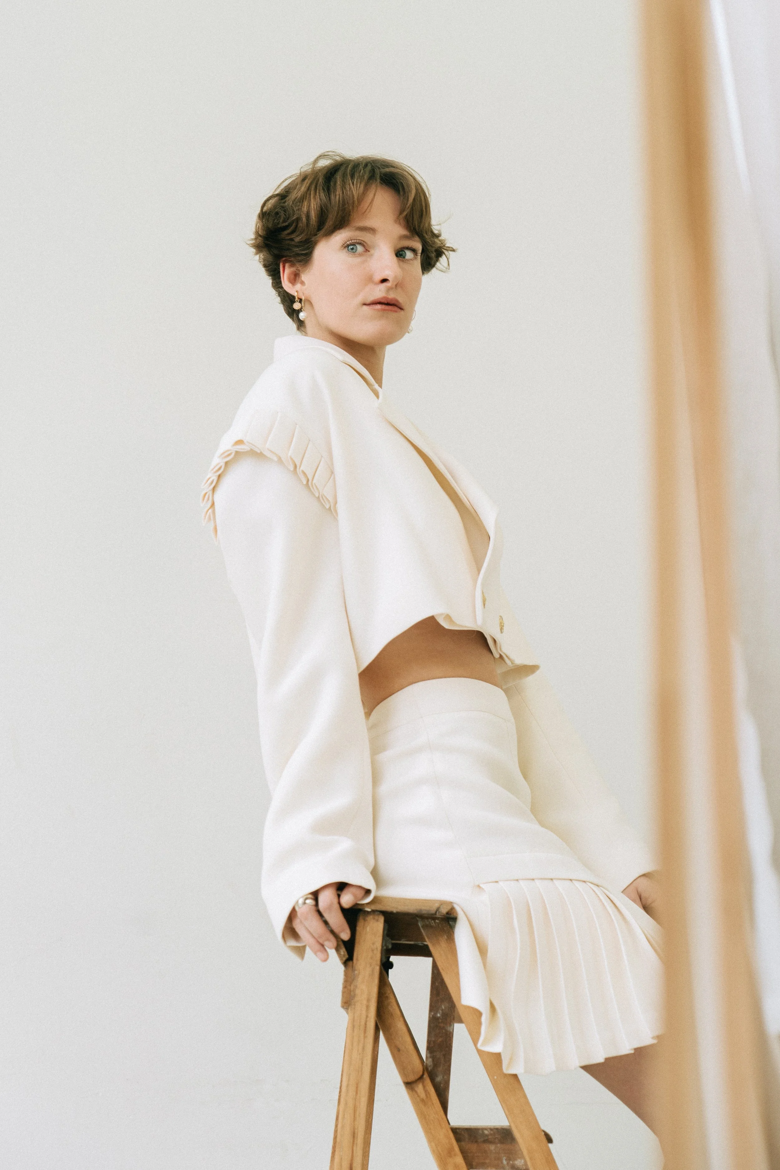 Une femme assise sur un escabeau en bois, portant une tenue blanche avec une jupe plissée et une veste courte, regardant vers la caméra dans un décor minimaliste.
