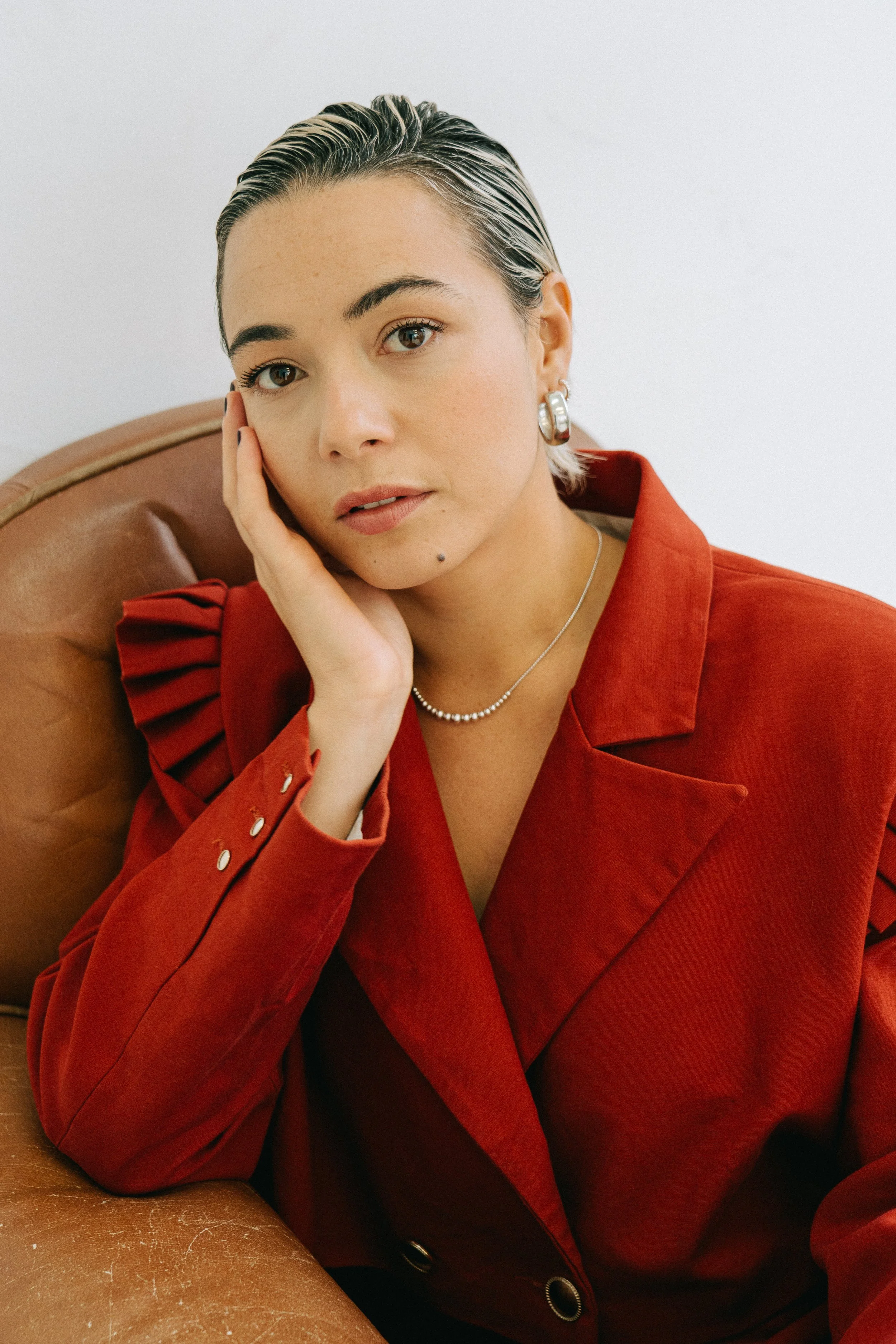 Une femme avec des cheveux courts et mouillés, portant une veste rouge, assise sur un fauteuil en cuir, avec un fond blanc.