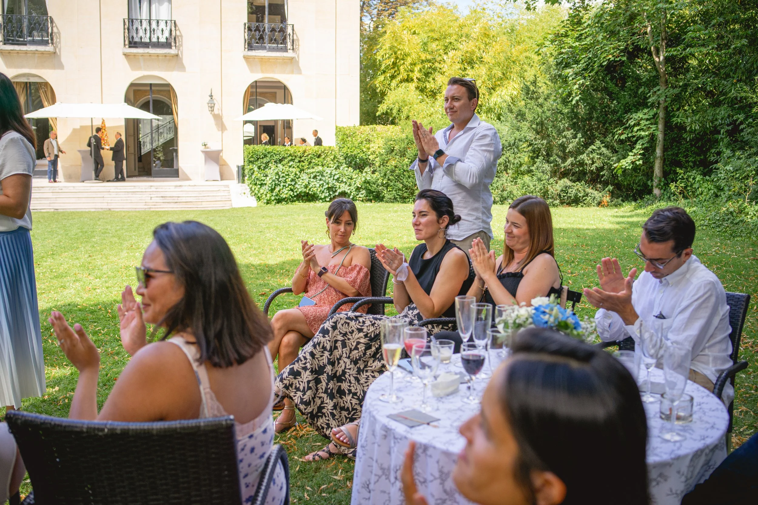 Groupe de personnes assises autour d'une table extérieure lors d'une fête ou d'une célébration, applaudi par un homme debout, avec un bâtiment en pierre et un jardin verdoyant en arrière-plan.