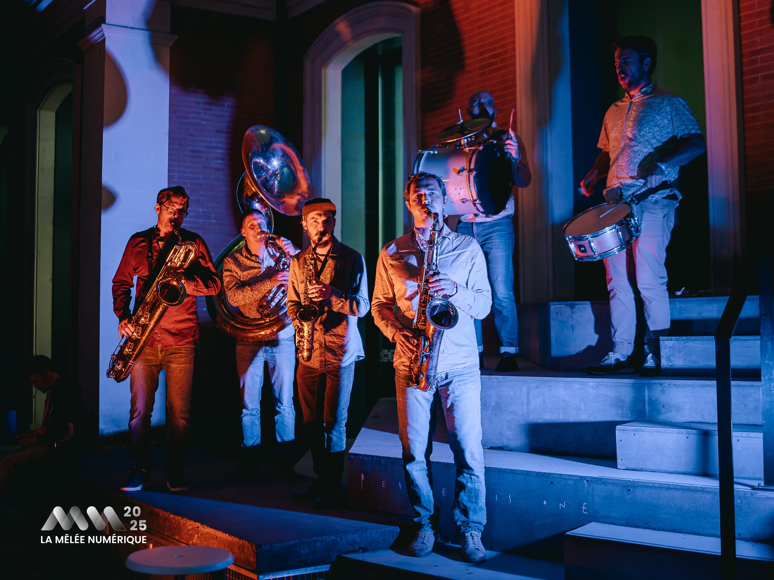 Un groupe de musiciens joue lors d'une performance en soirée. Ils sont équipés de divers instruments comme saxophone, trompette, batterie, et un clairon, et sont éclairés par des lumières colorées. Le logo '2025 La Mêlée Numérique' est visible en bas