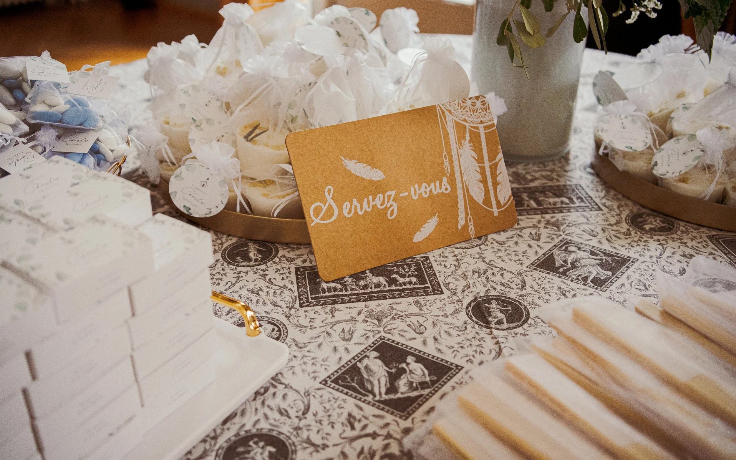 Table avec des petits cadeaux emballés, un panneau orange avec la phrase 'Servez-vous', et une nappe décorative. Il y a aussi une plante dans un pot blanc en arrière-plan.