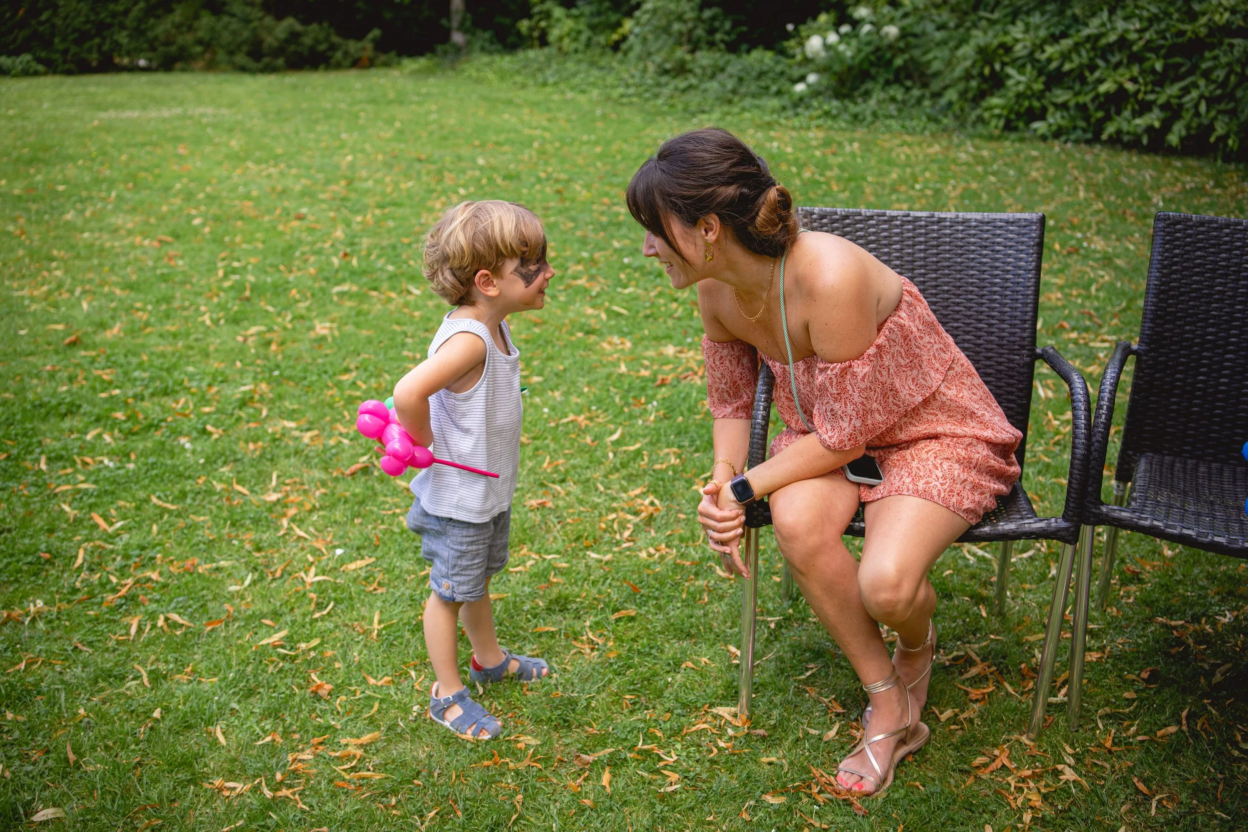 Une femme et un enfant en discussion dans un jardin. La femme est assise sur une chaise, la fille se tient debout devant elle, portant un ballon rose en forme de chien. Le garçon a le visage peint en face.