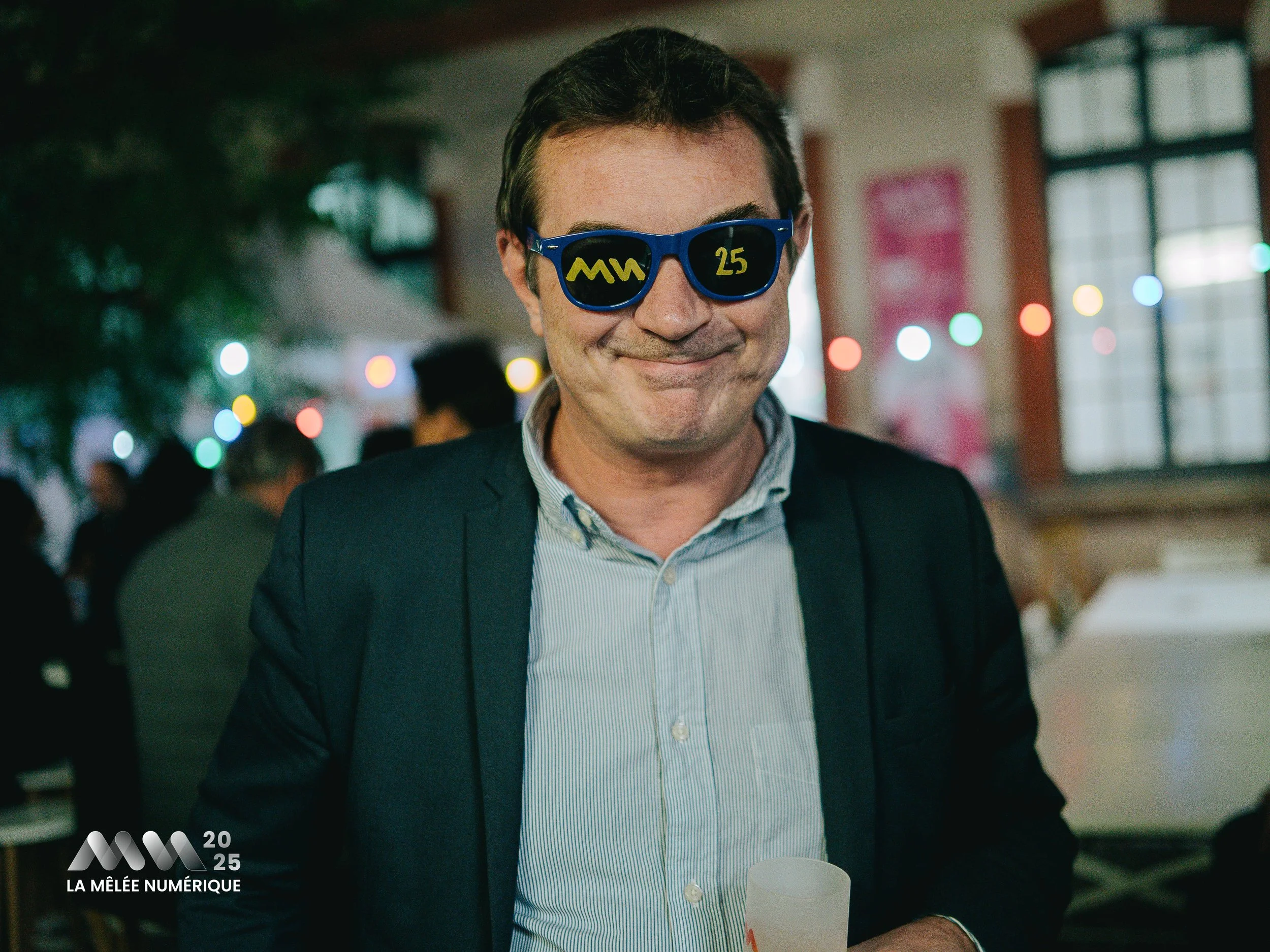 Homme souriant portant des lunettes de soleil avec le logo et l'année 2025, lors de l'événement La Mêlée Numérique 2025, dans une ambiance festive en intérieur.