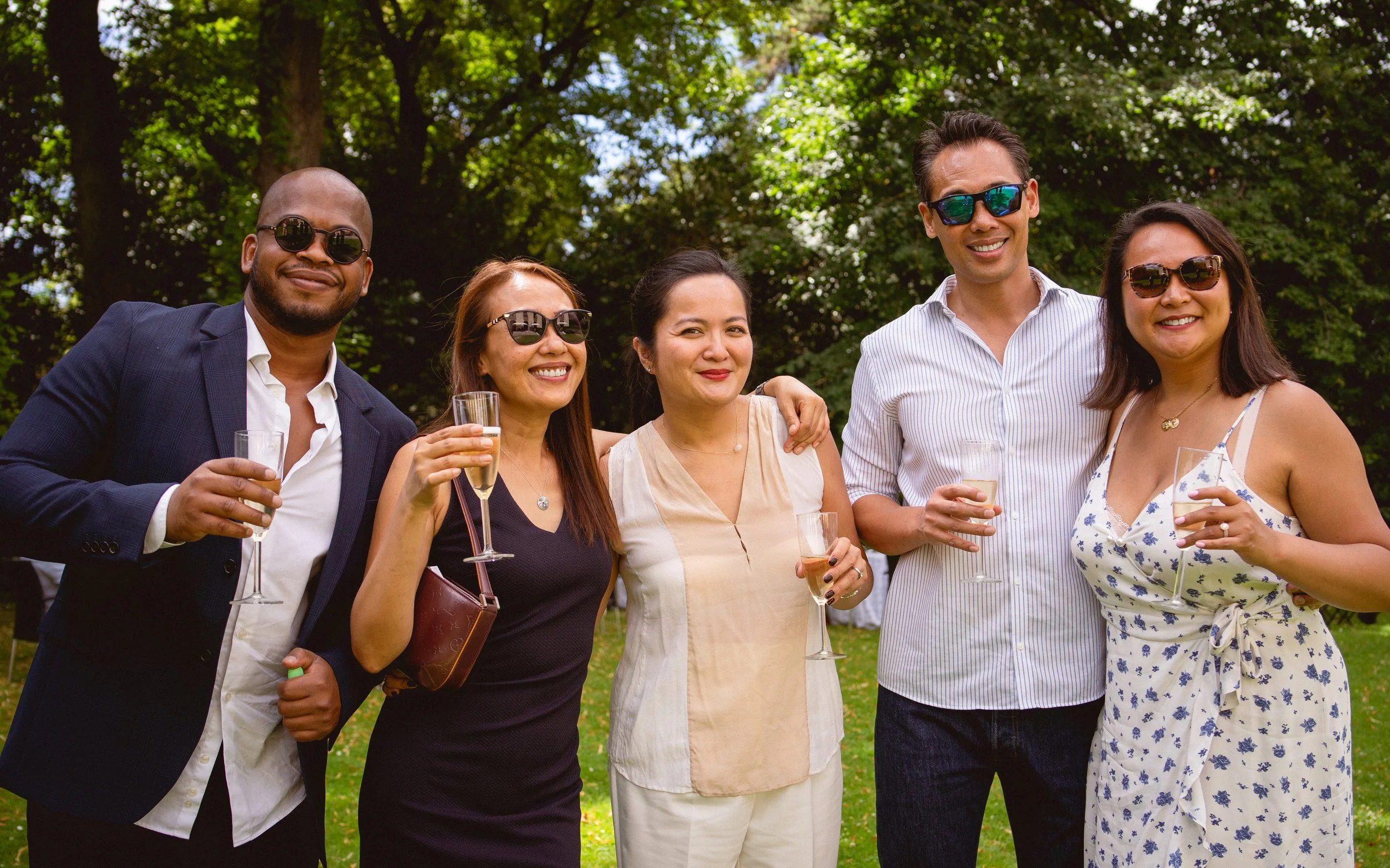 Groupe de six personnes souriantes avec des lunettes de soleil, tenant des verres de champagne, lors d'une fête en plein air dans un parc verdoyant en été.