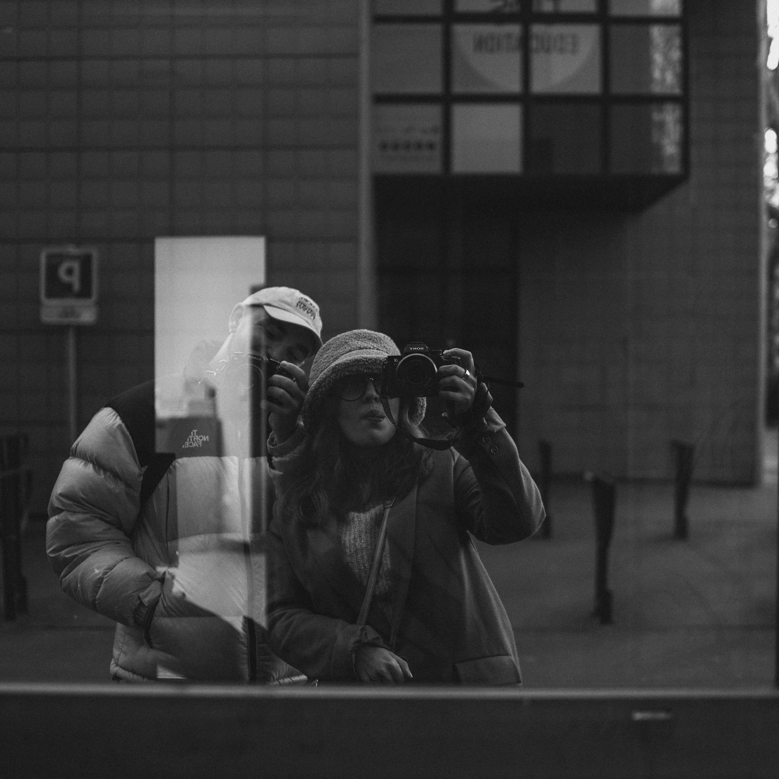 Deux personnes prennent un selfie dans une surface réfléchissante. La personne à l'avant porte un bonnet et une veste et tient un appareil photo, tandis que la personne derrière porte une casquette et un gilet. Le reflet montre un environnement urbai