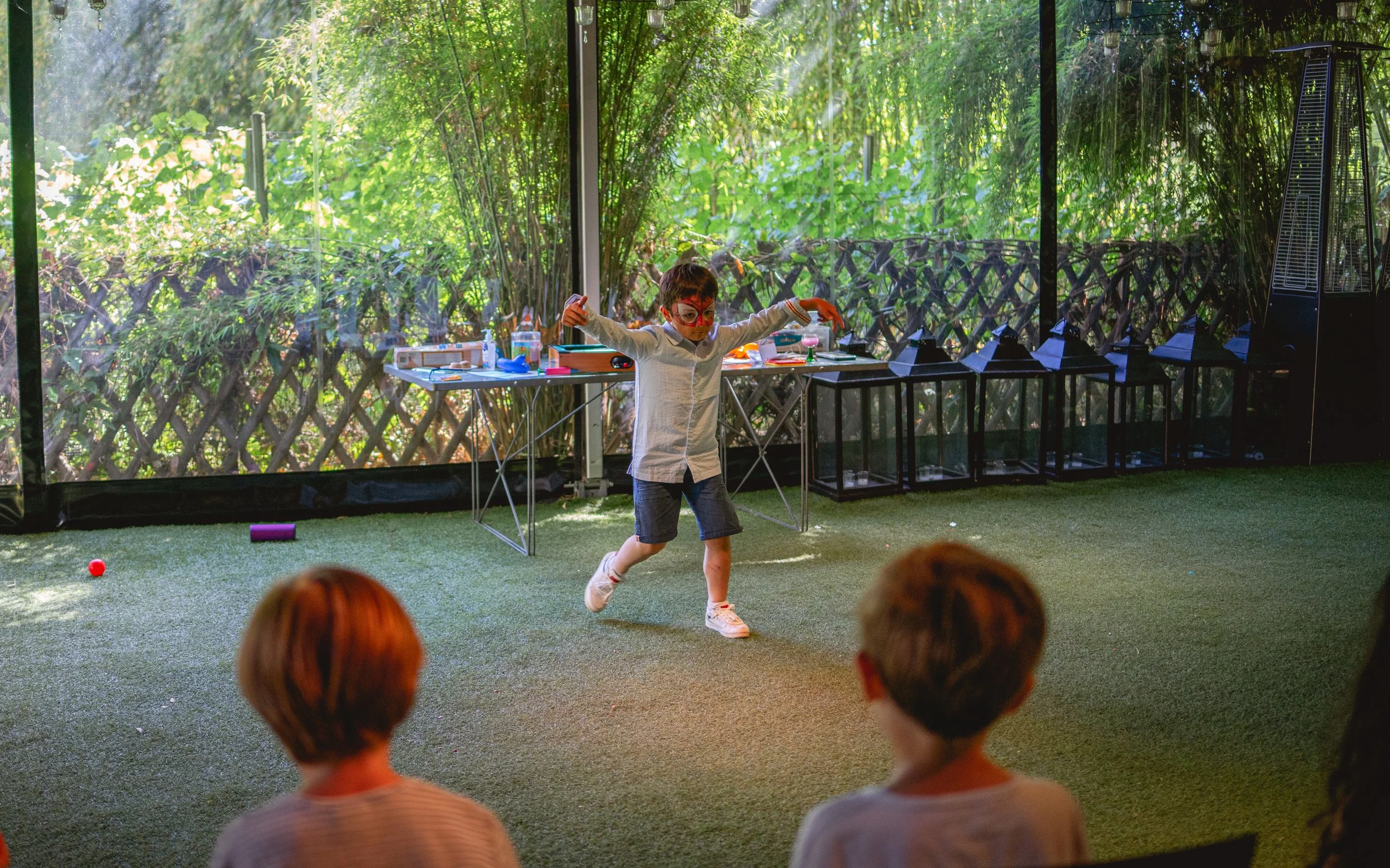 Un enfant avec un visage peint, jouant à une activité dans une salle avec de grandes fenêtres donnant sur un espace vert. Deux autres enfants regardent le jeu, assis face à l'enfant en mouvement.