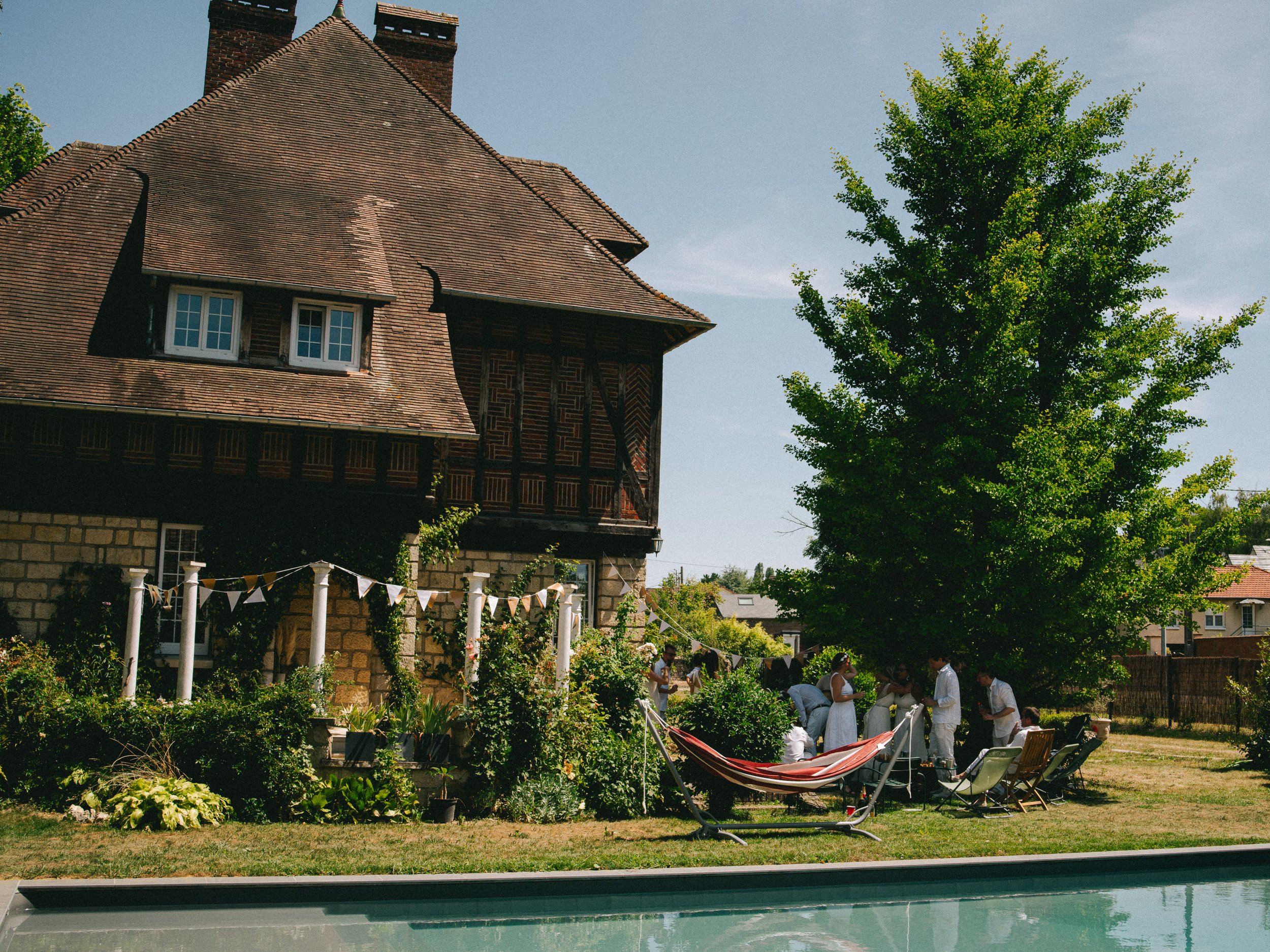 Une fête ou un rassemblement familial dans un jardin avec une maison ancienne, un grand arbre, une piscine, et des personnes en blanc autour d'une table.