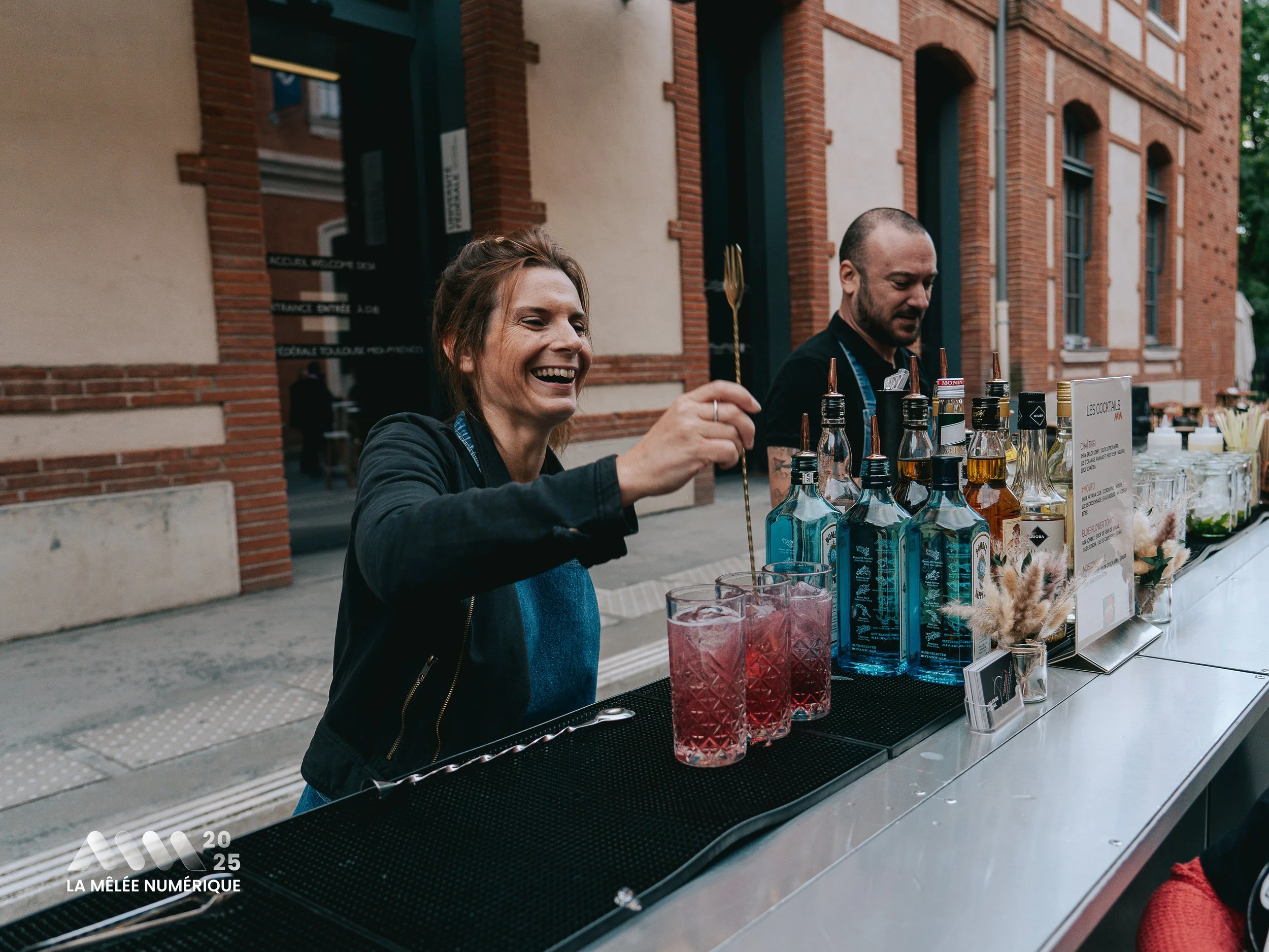 Une femme souriante prépare des cocktails derrière un stand en extérieur, avec un homme à ses côtés. Le stand affiche diverses bouteilles d'alcool et accessoires pour cocktails.